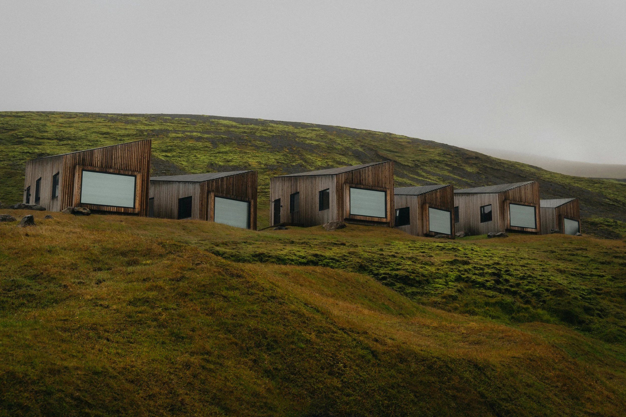 A series of modern, angular wooden houses with black-framed windows, situated on a grassy, slightly sloped landscape with a moss-covered hillside in the background under a gray, overcast sky.