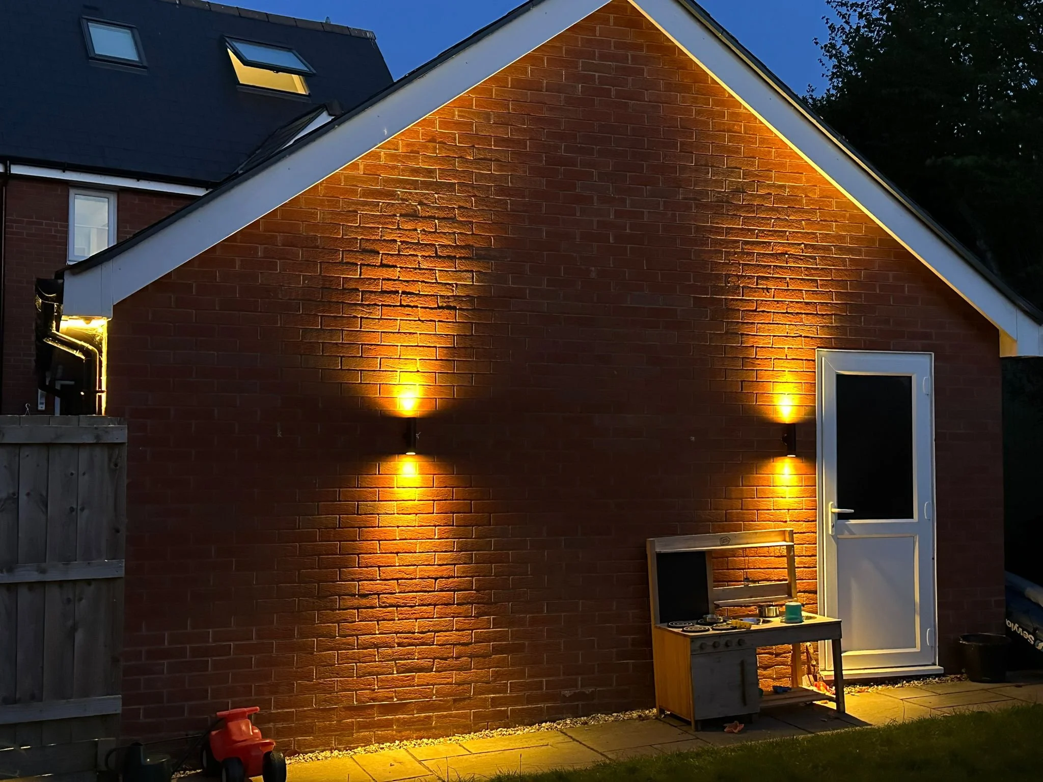 A brick house wall at night illuminated by outdoor lighting. The house has a pitched roof with windows. There is a small outdoor kitchen setup with a toy stove, a framed picture, and some small items on a table. A wooden fence is on the left, and a door with glass at the bottom right of the image.