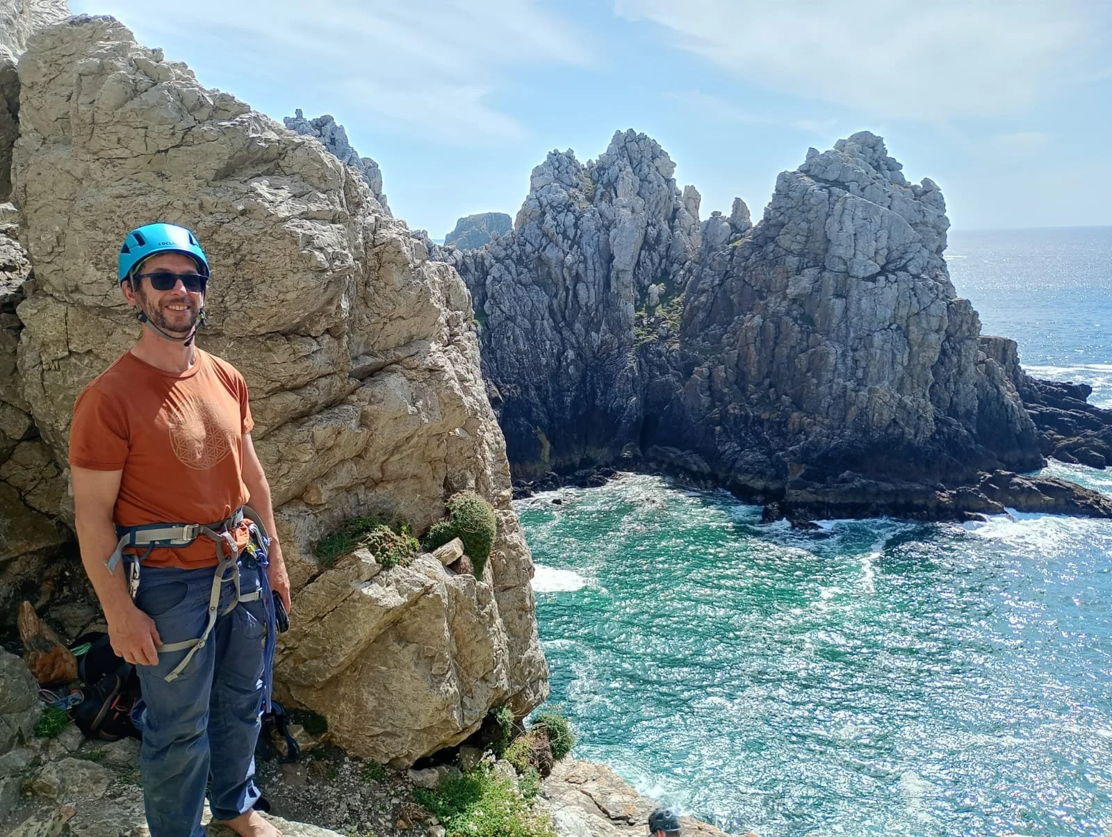 A man in outdoor clothing and a blue helmet standing on rocky cliffs by the ocean, with large rock formations and blue waters in the background.