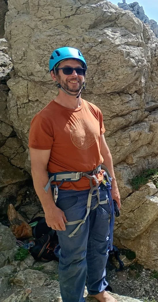 A man wearing a blue helmet, sunglasses, and climbing gear, standing outdoors next to a large rocky formation, smiling at the camera.