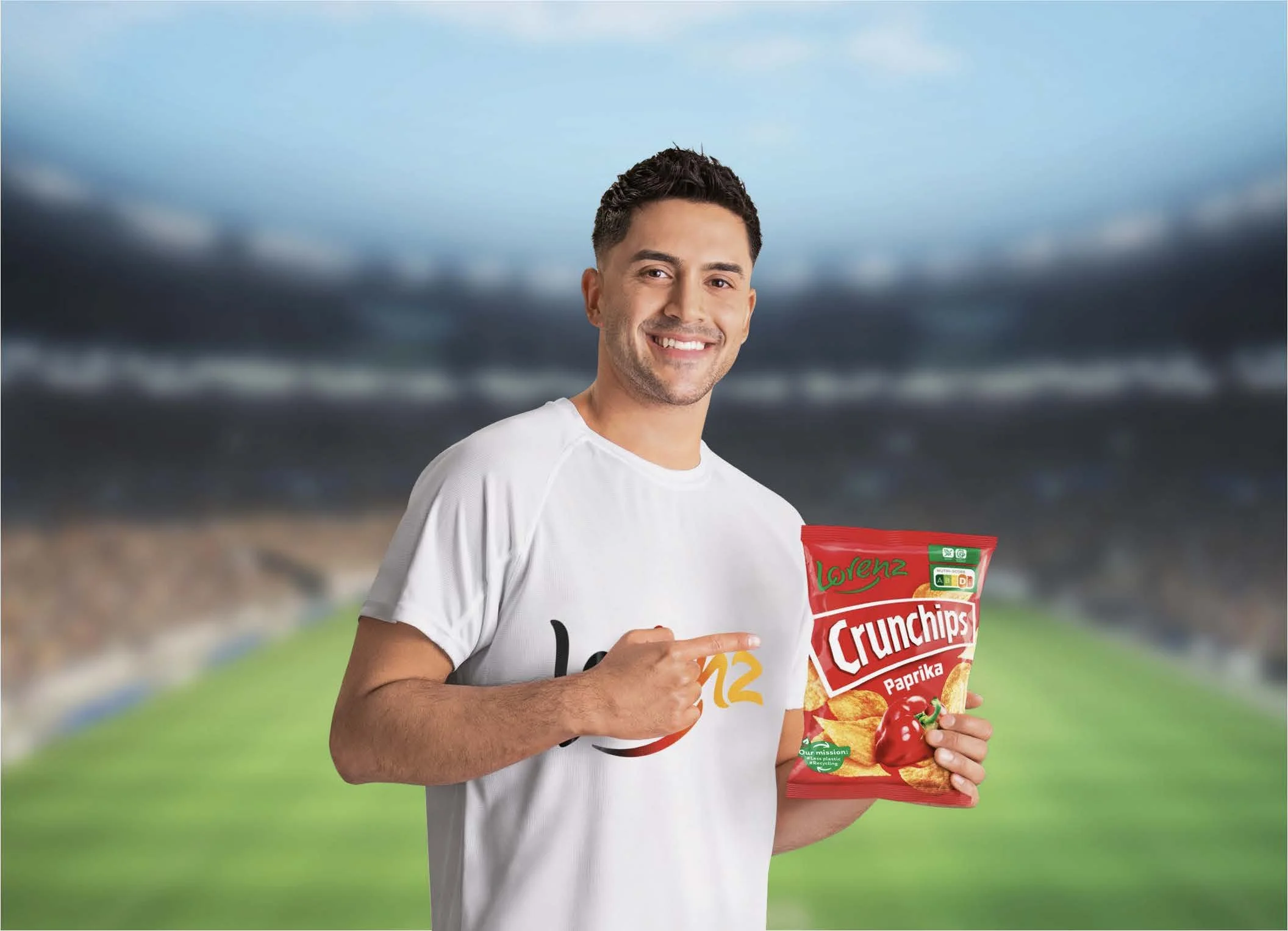 Ein lachender Mann in einem weißen T-Shirt hält eine Tüte mit Chips in der Hand. Der Hintergrund zeigt eine bewölkte Landschaft mit einem Sportstadion.