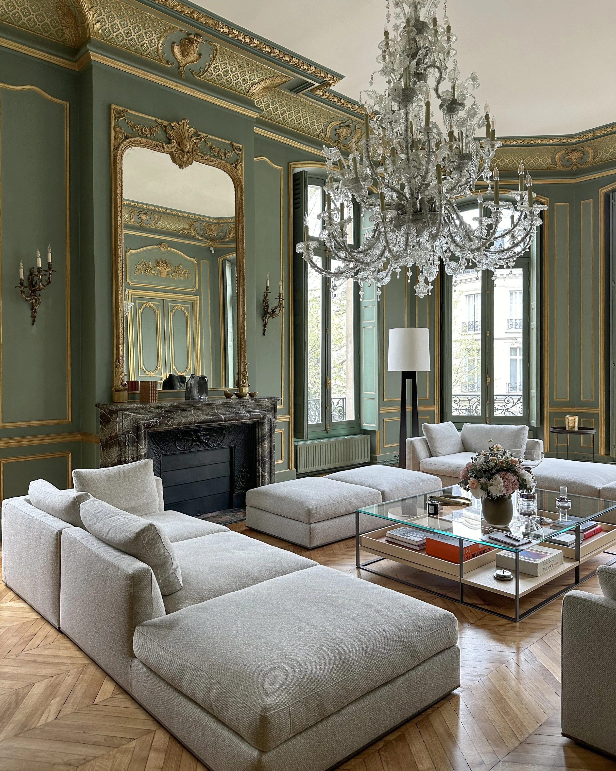 Salon élégant avec murs verts et moulures dorées, grand miroir, cheminée en marbre, chandelier en cristal, canapés blancs, table basse avec livres et fleurs, fenêtres avec vue urbaine.