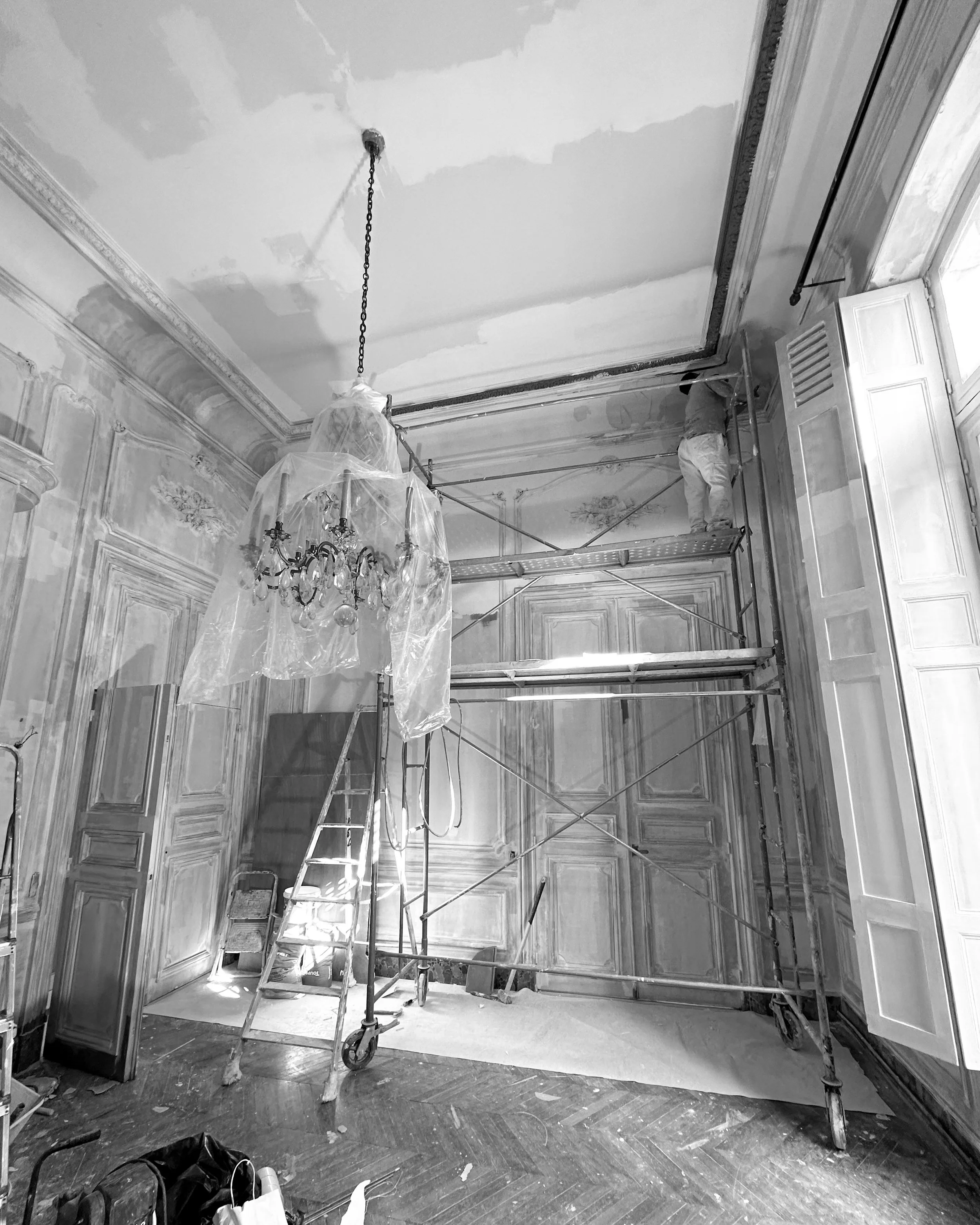 Une pièce en cours de rénovation avec escabeau, échelle, et un peintre travaillant sur le plafond. Le mobilier et la décoration sont en partie protégés par des plastiques.