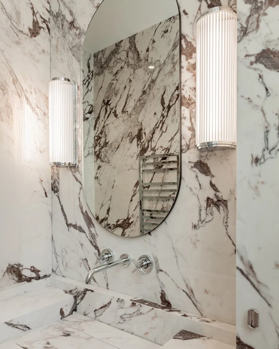 Un mur en marbre avec un miroir ovale, deux appliques murales en haut, un robinet suspendu et un évier en marbre dans une salle de bain élégante.