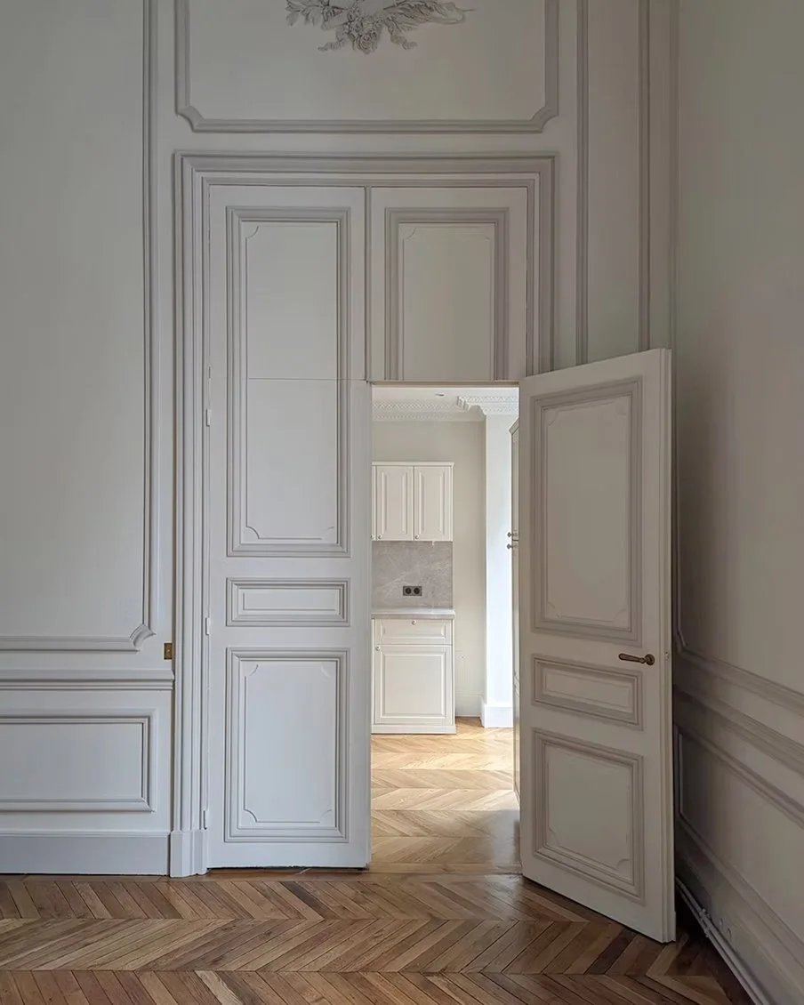 Vue intérieure d'une pièce avec des murs en bois peint blanc, un parquet en bois clair et une porte en bois blanc entrouverte menant à une cuisine.
