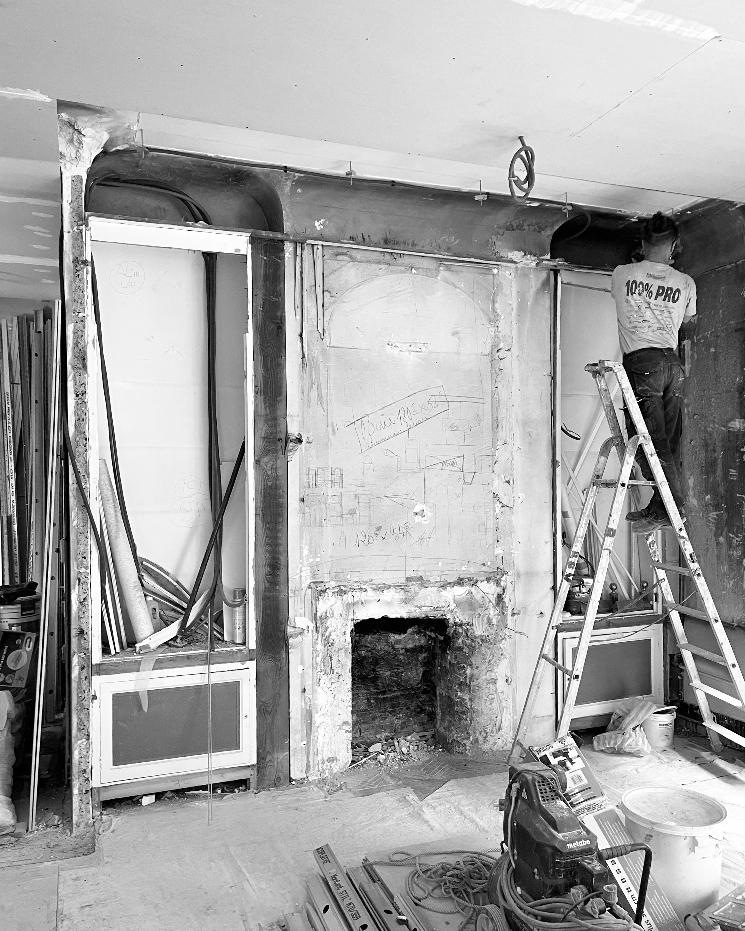 Travaux de rénovation intérieure avec un ouvrier en haut d'une échelle, murs en cours de démolition, outils et matériaux de construction au sol.