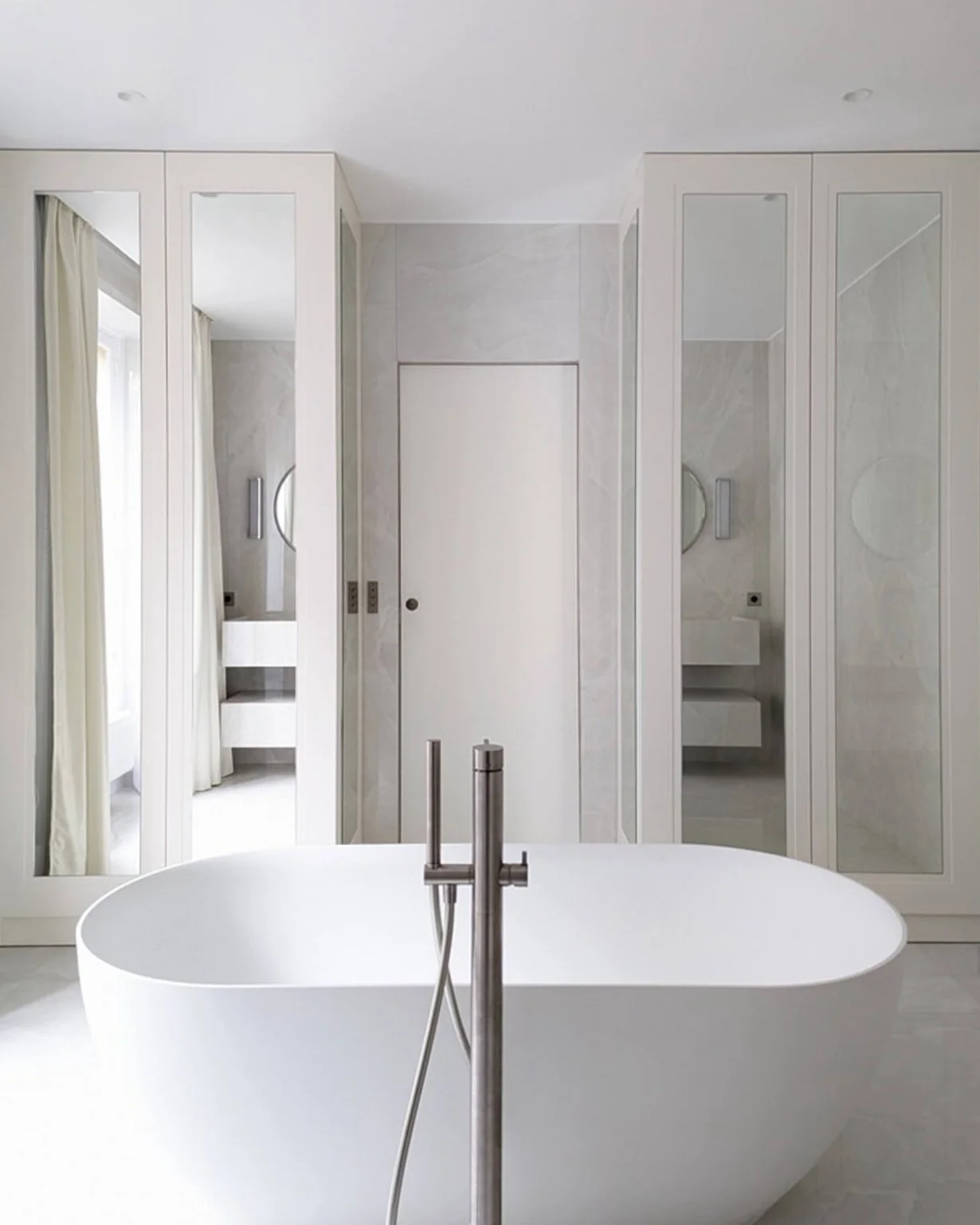 Une salle de bain moderne avec une baignoire blanche ovale, un sol clair, des armoires hautes avec des miroirs et un lavabo blanc. La pièce est lumineuse avec des fenêtres à rideaux blancs.