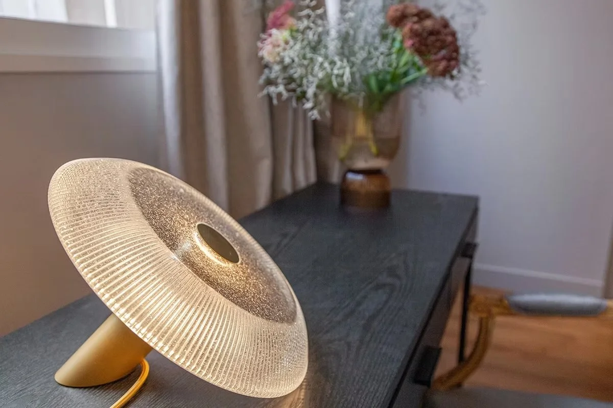 Lampe de bureau moderne avec un abat-jour en verre textured sur une table en bois, avec un vase de fleurs en fond dans une pièce illuminée.