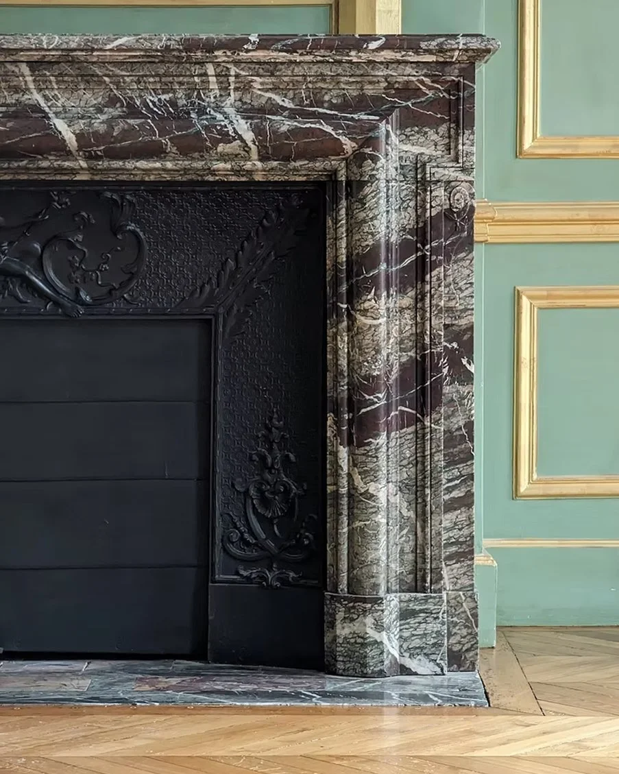 Cheminée en marbre vert foncé avec détails sculptés en noir, dans une pièce avec murs verts et moulures dorées.