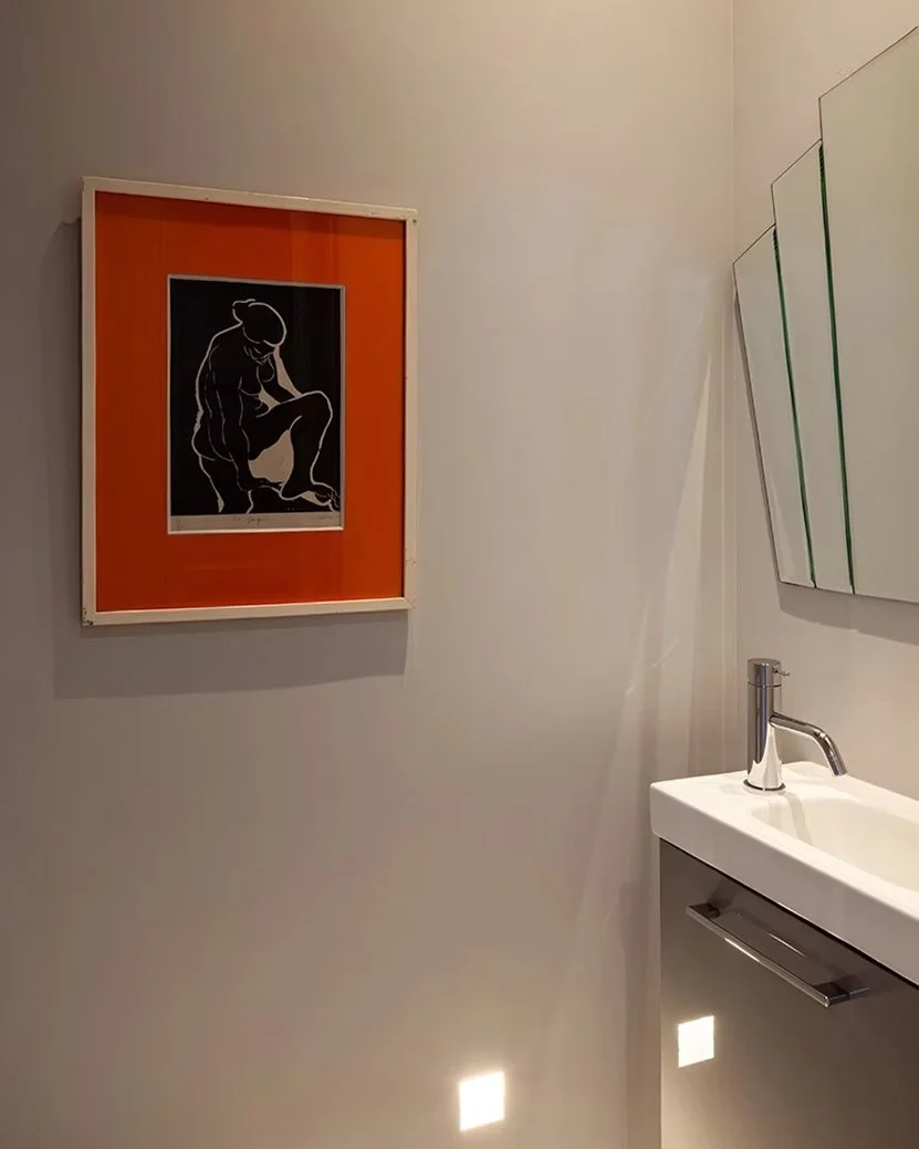 Une salle de bain minimaliste avec un tableau moderne représentant une silhouette en noir et blanc sur un fond orange, un lavabo blanc avec un robinet métallique, et un miroir abstracte avec des lignes vertes.