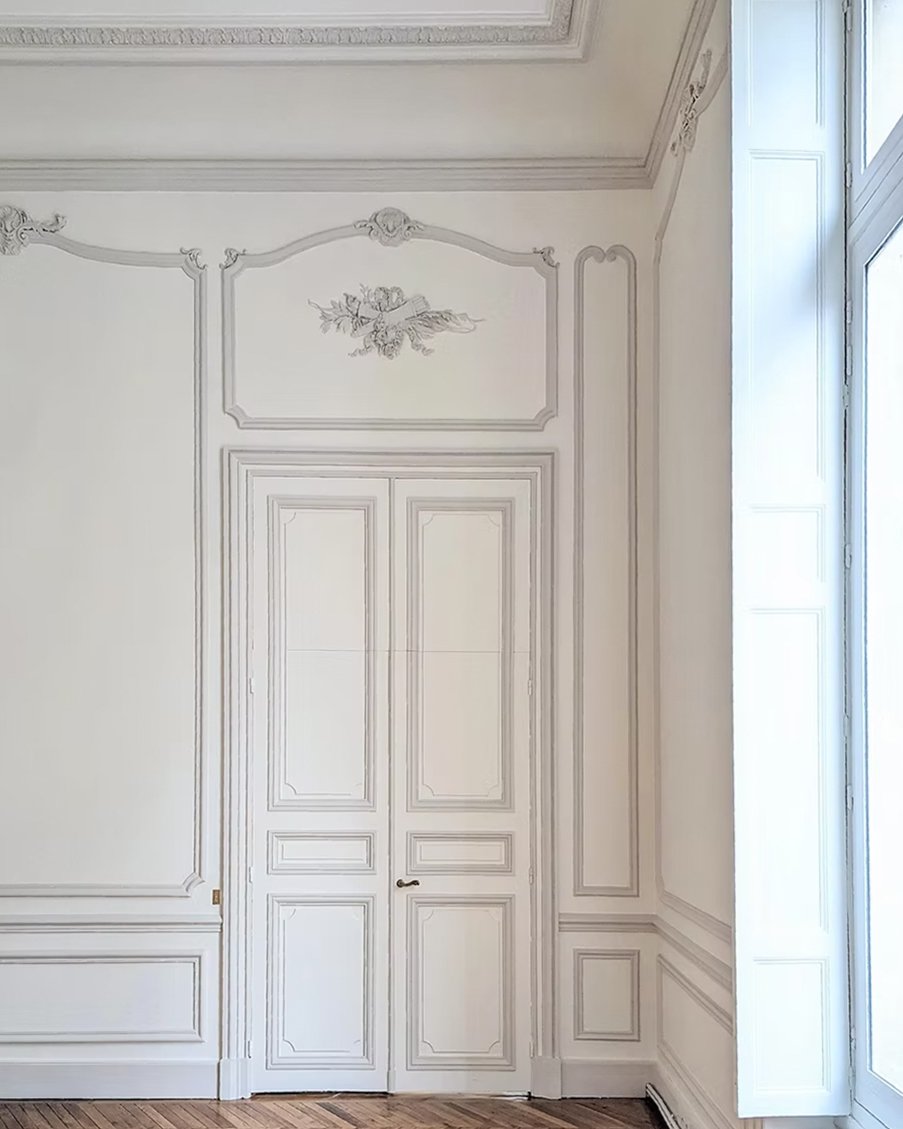 Une pièce avec des murs et portes blancs ornés de moulures décoratives, un plafond mouluré et un parquet en bois, lumière naturelle entrant par une grande fenêtre.