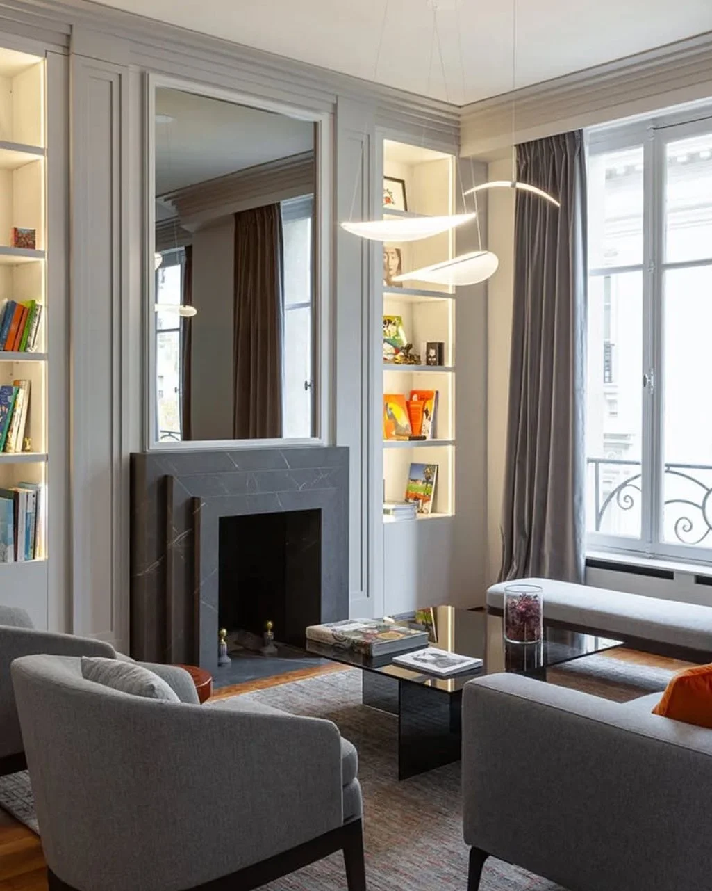 Salon contemporain avec cheminée en marbre, bibliothèque, canapé gris, table basse noire, grand windows avec rideaux gris, suspension moderne, décorations et livres.