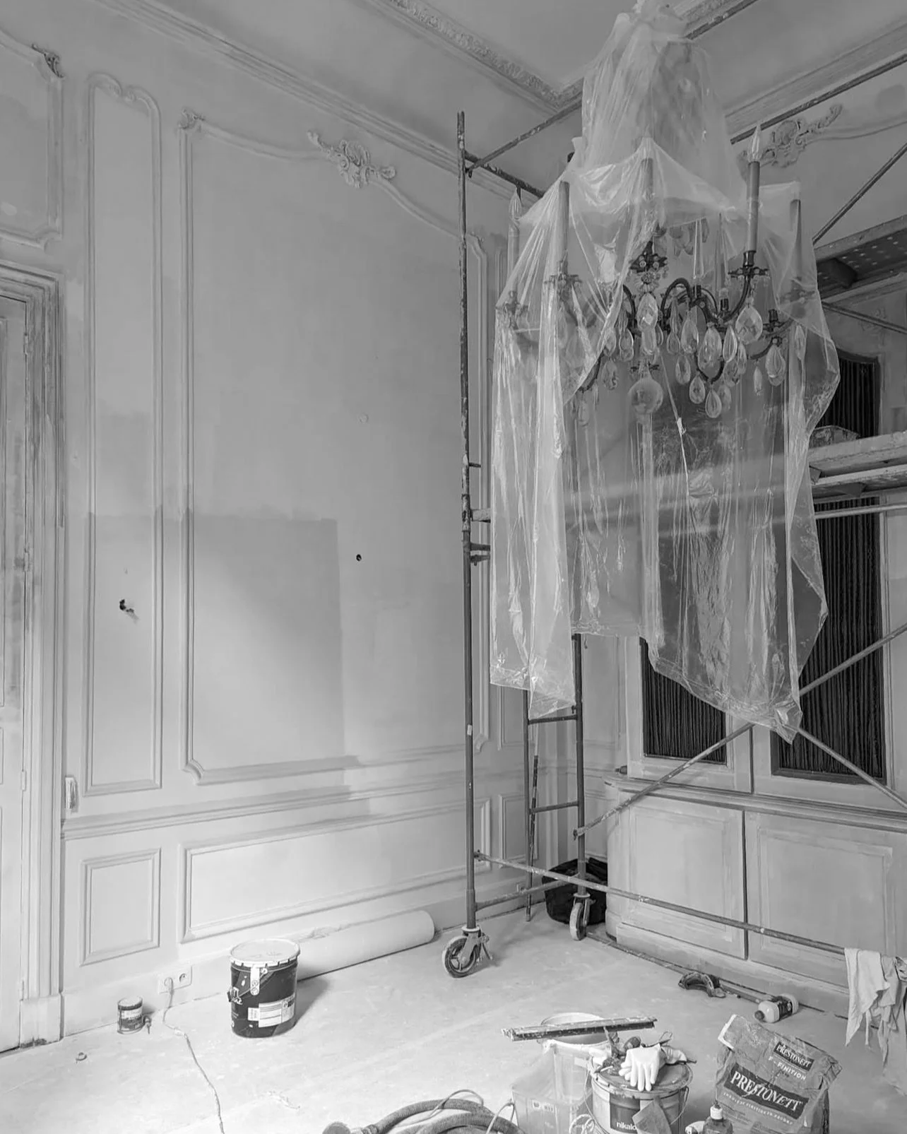 Chantier de rénovation intérieur avec un échafaudage, un lustre et des outils, dans une pièce avec moulures au plafond et panneaux en bois.