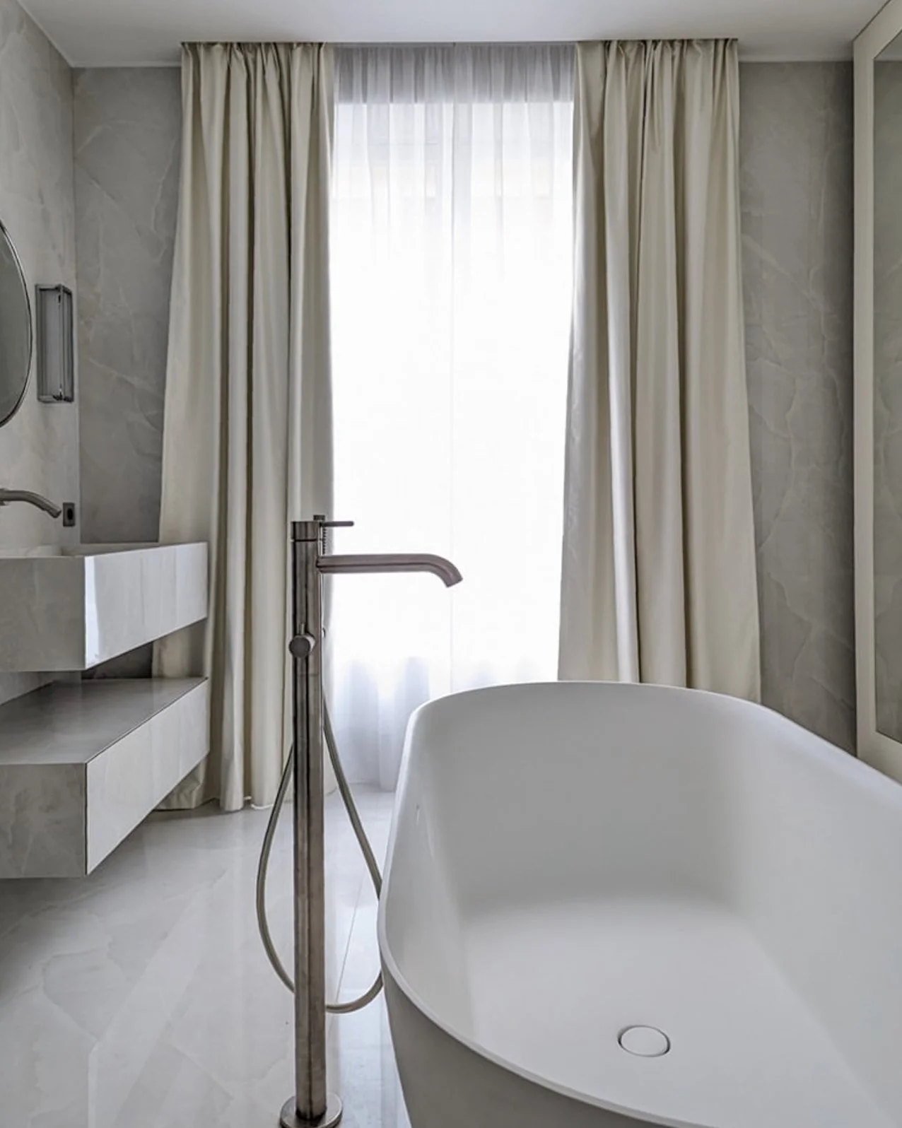 Une salle de bain avec une baignoire en forme ovale, un robinet de baignoire, un lavabo blanc avec un miroir, des rideaux blancs devant une grande fenêtre laissant entrer la lumière naturelle.