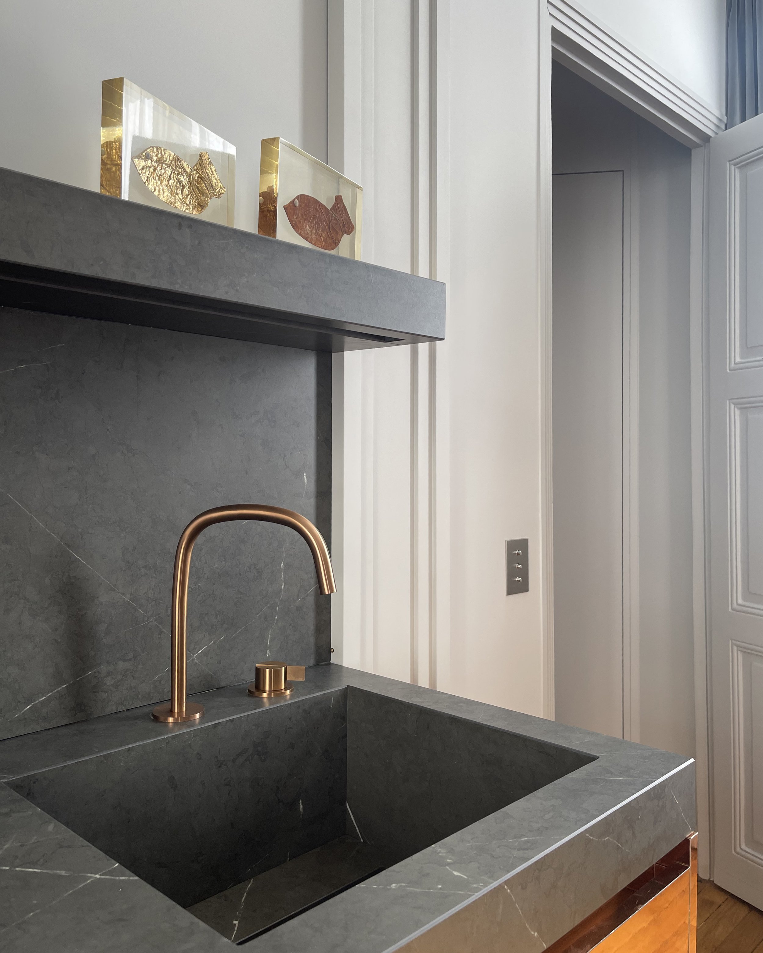 Un évier en pierre grise avec un robinet en cuivre et deux cadres muraux avec des poissons en or et en cuivre au-dessus, dans une pièce avec murs blancs et porte blanche avec moulures.