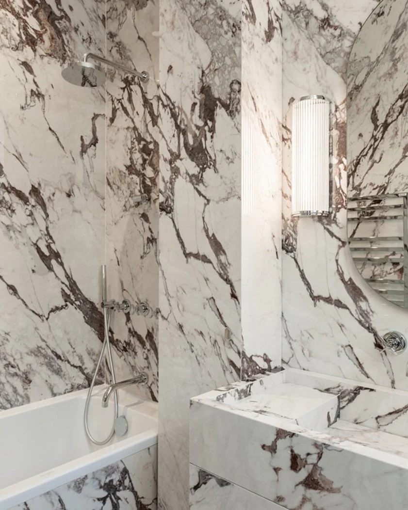 Salle de bain avec murs en marbre blanc et brun, douche à gauche, lavabo en marbre à droite, et miroir rond. Éclairage mural à droite.