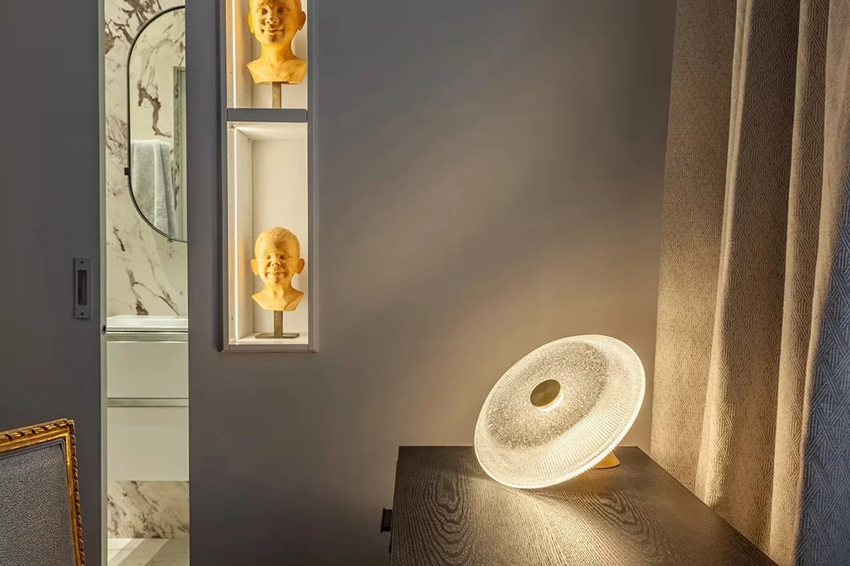 Une pièce blanche avec un miroir ovale, une étagère avec deux bustes en plâtre jaunes, une lampe de bureau moderne sur une table en bois, et un rideau beige à motif.