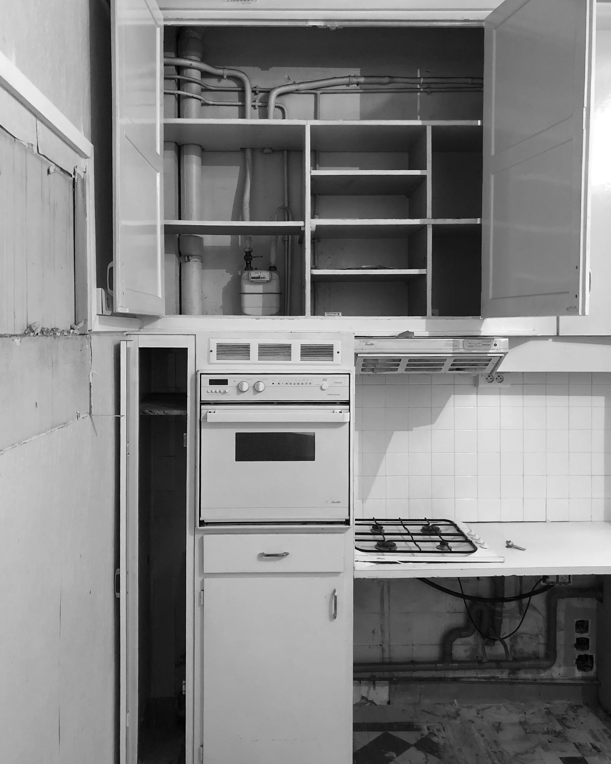 Cuisine avec four, plaques à gaz, placards vides et tuyaux visibles, en noir et blanc.
