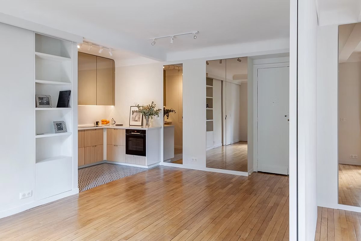 Intérieur d'un appartement moderne avec cuisine ouverte, plancher en bois naturel, étagères blanches, miroir mural, et décoration minimaliste.