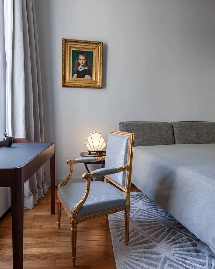 Intérieur d'une chambre avec une peinture de portrait d'une jeune fille dans un cadre doré, un fauteuil ancien en bois avec rembourrage bleu, une lampe moderne, un lit gris et un bureau avec des rideaux gris à gauche.