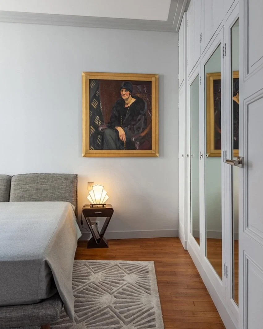 Une chambre avec un lit à gauche, une lampe de table allumée à côté du lit, un tableau représentant une femme assise encadré en bois au mur, des armoires blanches avec des miroirs sur la droite et un tapis beige avec un motif géométrique au sol.