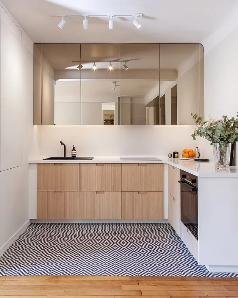 Cuisine moderne avec placards en bois clair, plan de travail blanc, grand miroir récent au mur, tapis à motifs géométriques noirs et blancs, éléments décoratifs comme des citrons, un vase avec des feuilles, et différents appareils électroménagers.