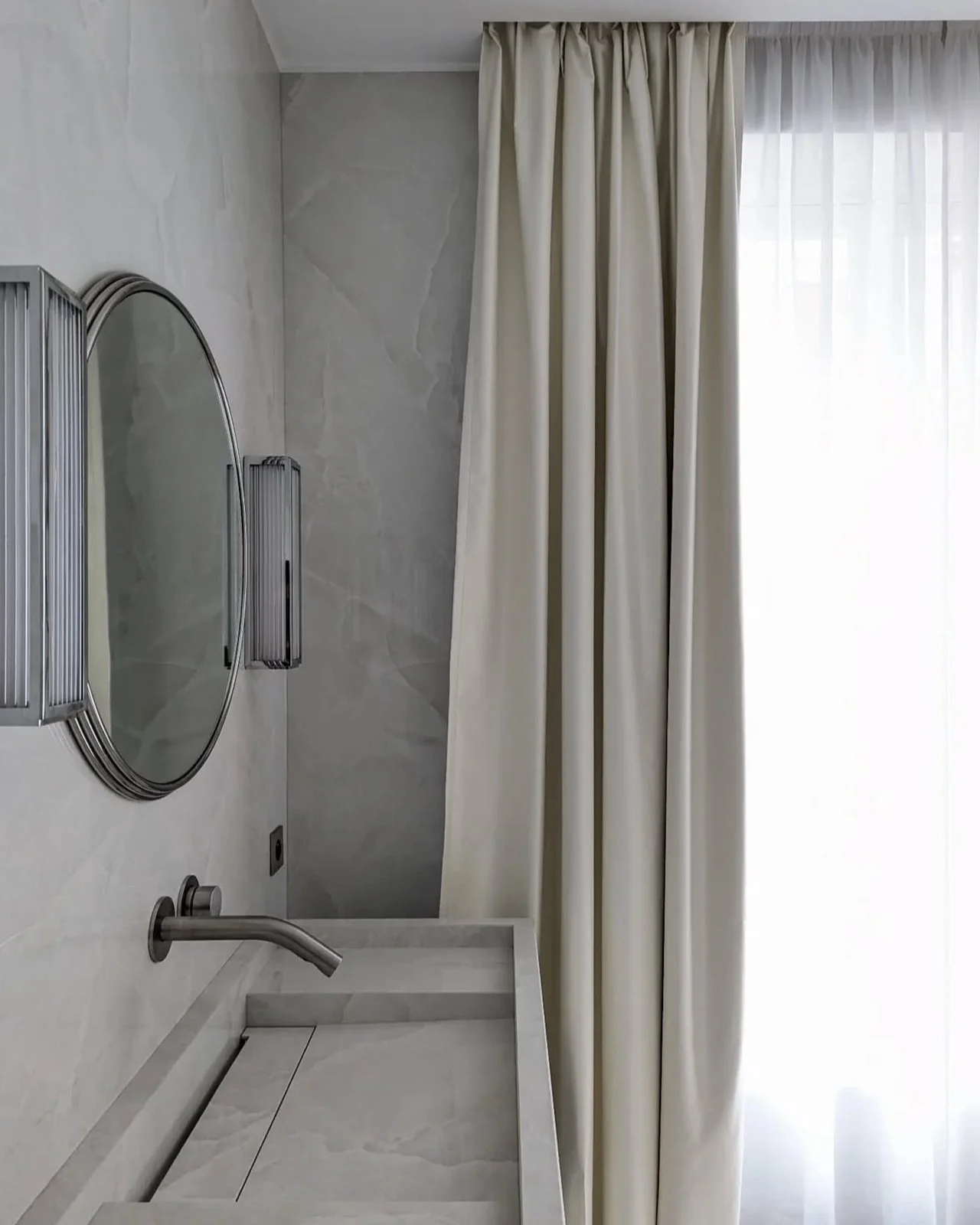 Intérieur d'une salle de bain avec un miroir rond, un évier en marbre, un robinet en métal, des murs en carrelage clair, des rideaux en tissu clair, et une fenêtre avec lumière naturelle.