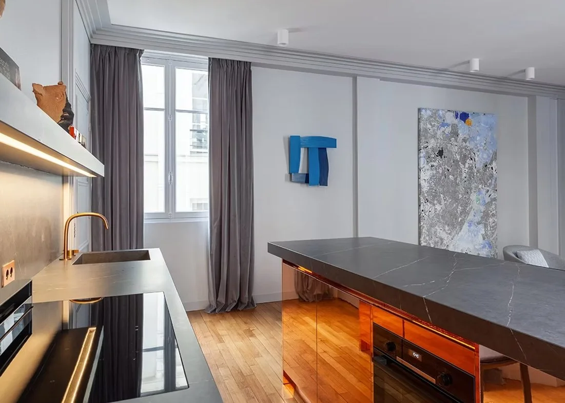 Une cuisine moderne avec un comptoir en marbre noir, un évier en inox avec un robinet doré, des planches en bois clair, des rideaux gris, une fenêtre, des œuvres d'art sur le mur et un sol en bois.