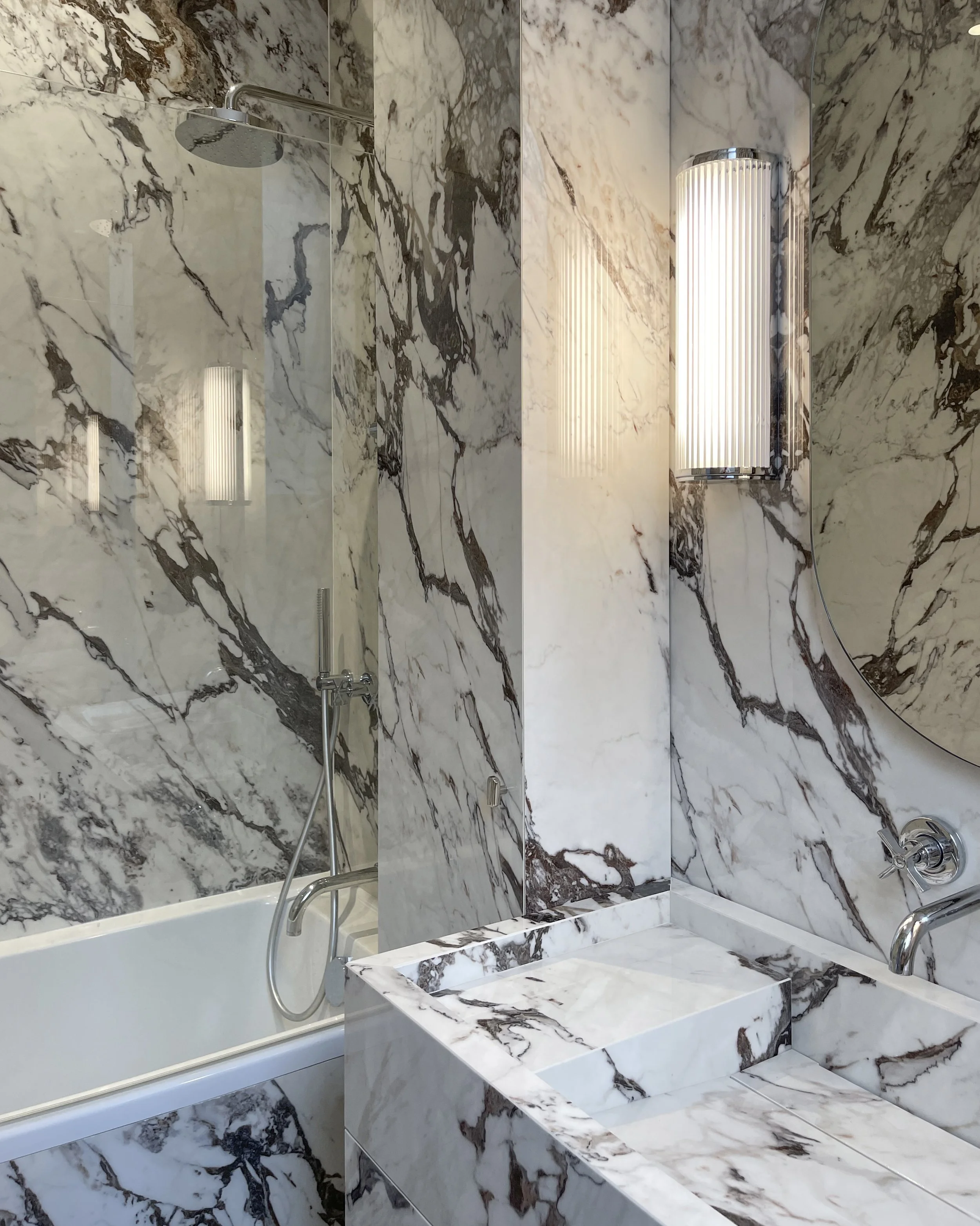 Salle de bain en marbre blanc avec veines noires, douche à l'italienne, lavabo en marbre, miroir rond et luminaire mural moderne.