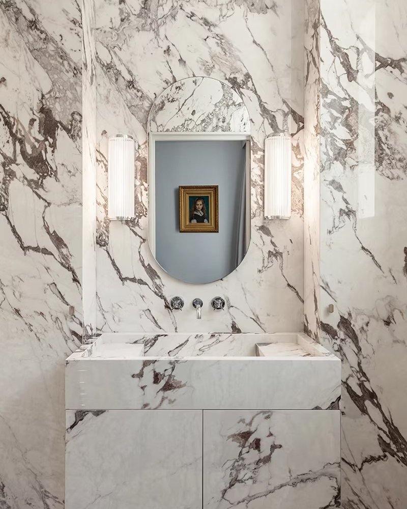 Un lavabo en marbre avec un miroir ovale au-dessus, deux appliques murales lumineuses à gauche et à droite, et un tableau encadré derrière le miroir.