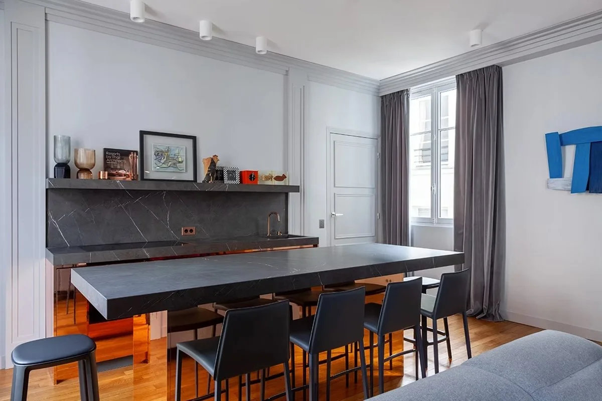 Salle à manger ou cuisine avec un grand plan de travail en marbre noir, une table en marbre noir, plusieurs chaises noires, une fenêtre avec rideaux gris, décoration moderne sur le mur et des objets décoratifs sur le mur.
