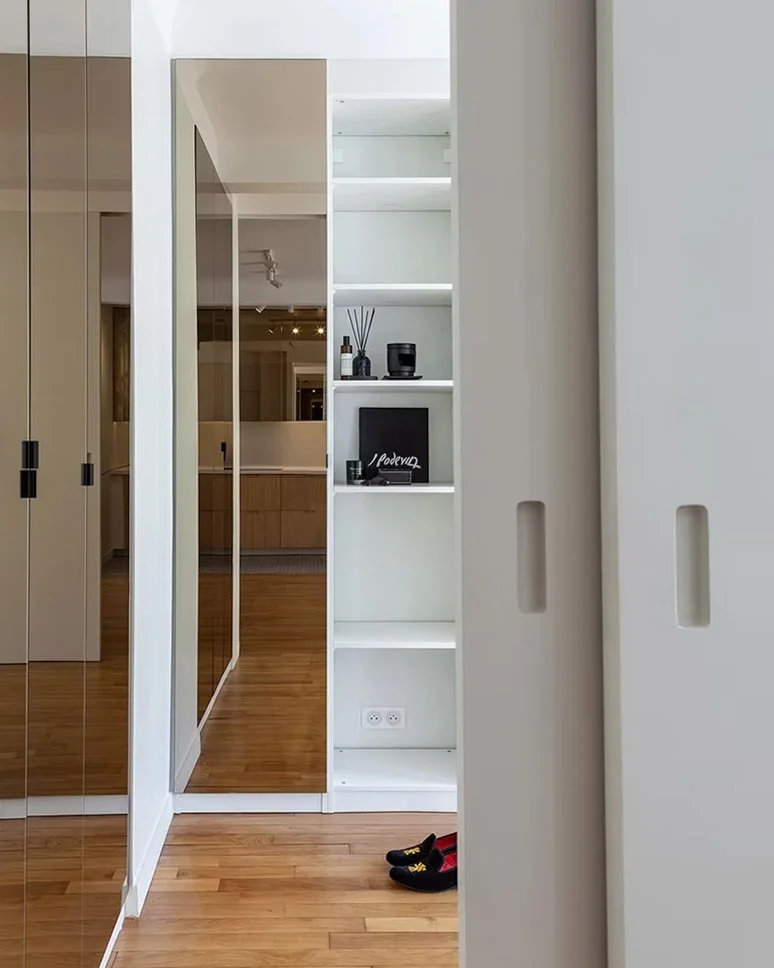 Vue d'un intérieur moderne avec une étagère blanche contenant des objets décoratifs, un miroir, un sol en bois clair, et une paire de chaussures noires et rouges posées au sol.