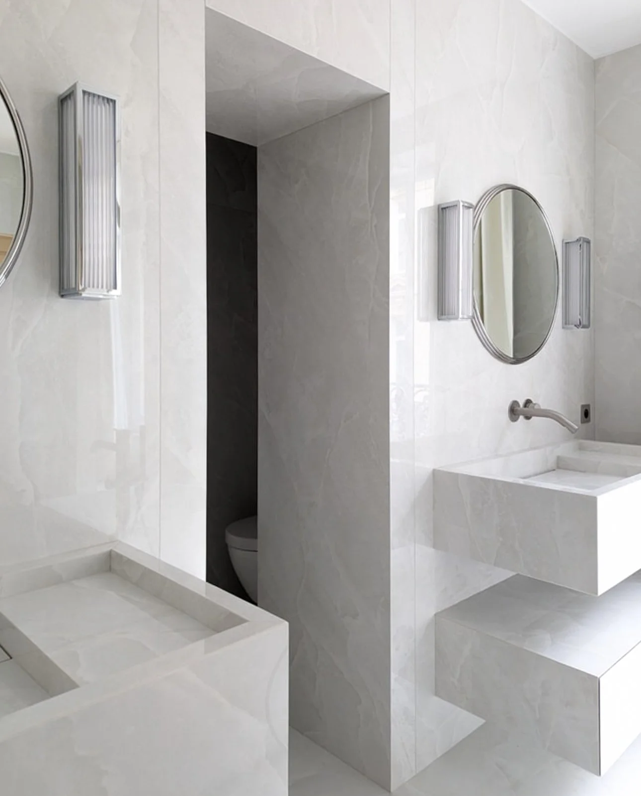 Salle de bain moderne avec murs en marbre blanc, miroir oval, lavabo à angle droit, et éclairages muraux.