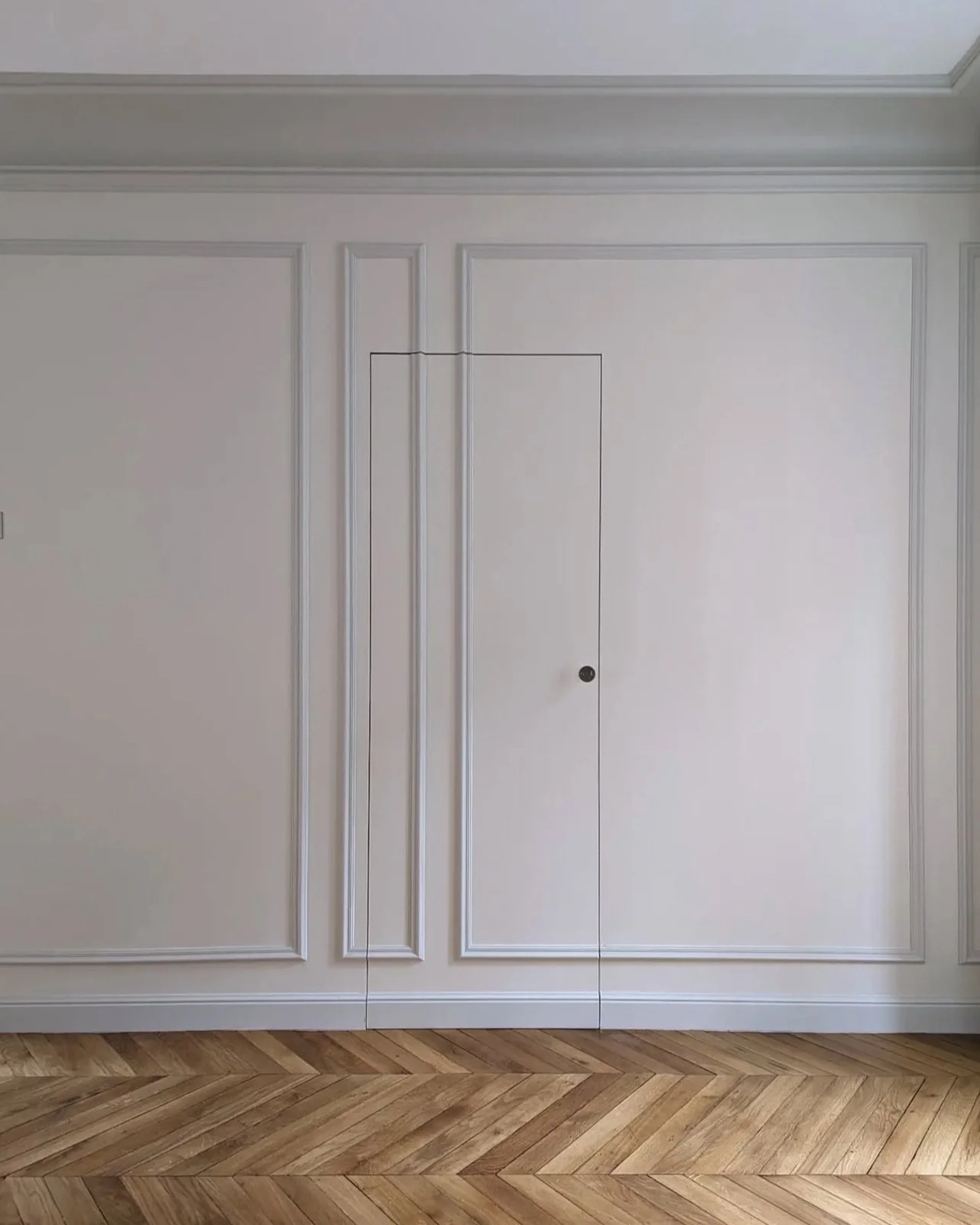 Mur blanc avec moulures et porte cachée dans le mur, plancher en parquet en diagonale.