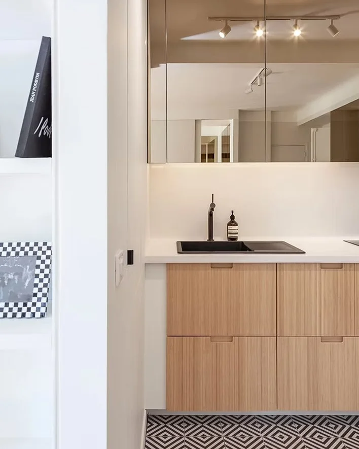 Coin cuisine avec un évier noir, meuble en bois clair, miroir mural, plafond à spots, et sol à motifs géométriques noir et blanc.