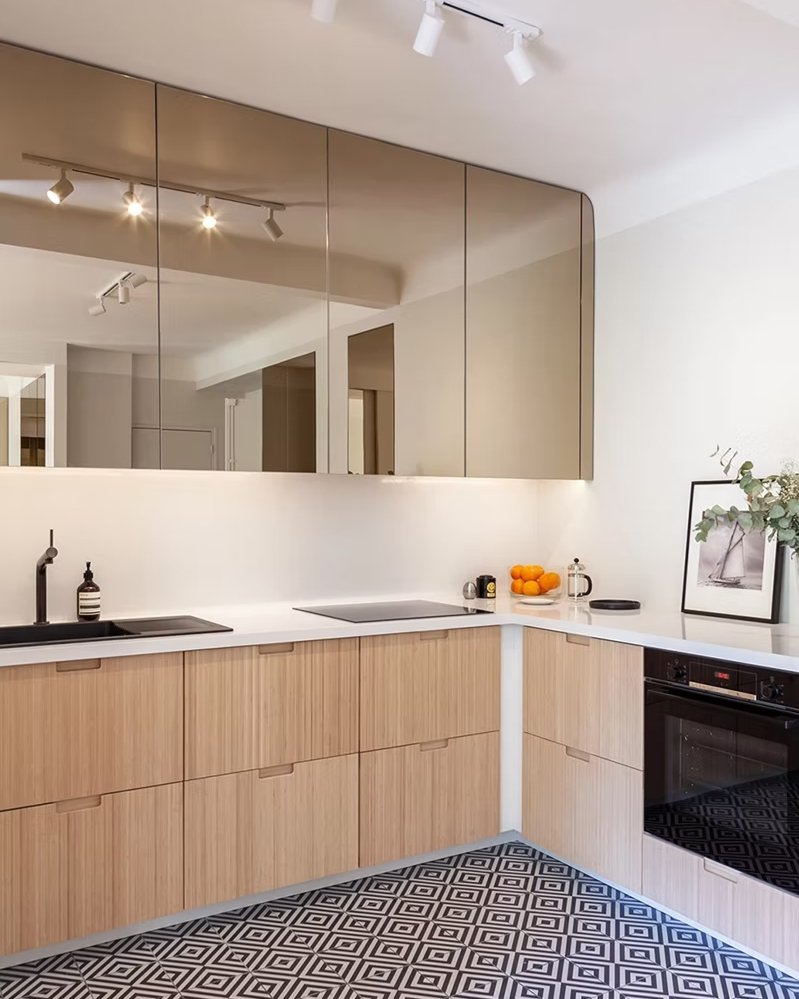 Cuisine moderne avec des façades en bois clair, un évier noir, une plaque de cuisson, un four noir, un miroir mural, des objets décoratifs, et un plan de travail blanc.