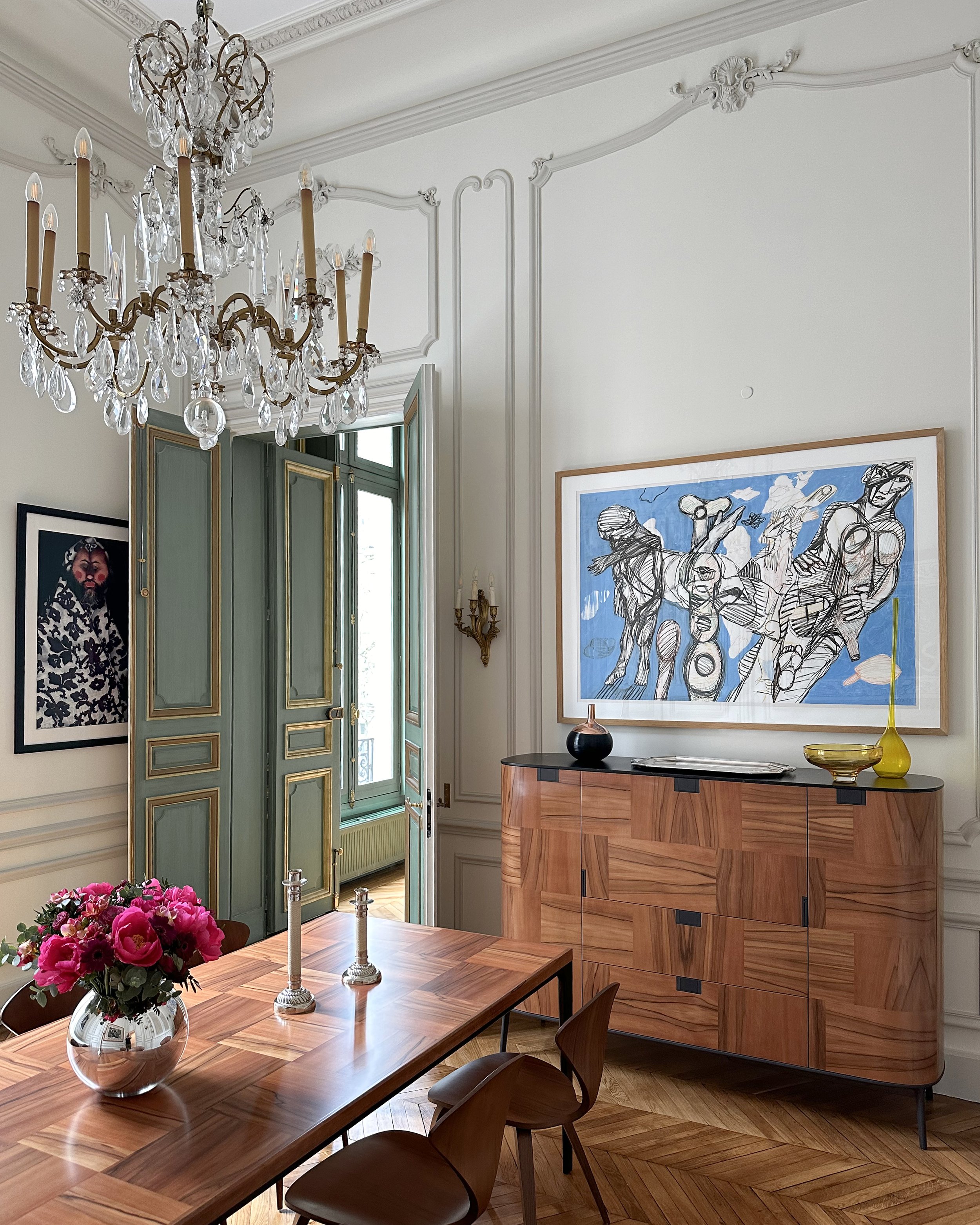 Une salle à manger élégante avec une table en bois, un lustre en cristal suspendu, un tableau moderne accroché au mur, et des meubles en bois avec décorations comme des vases et des bougies.