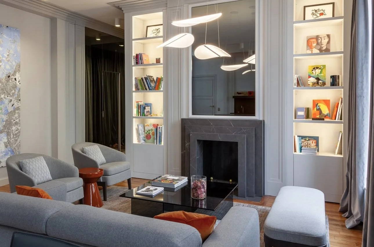 Salon avec cheminée en marbre noir, étagères intégrées avec des livres, deux fauteuils gris avec coussins, un canapé gris, un tapis, un tableau mural abstrait, et un lampadaire suspendu moderne.