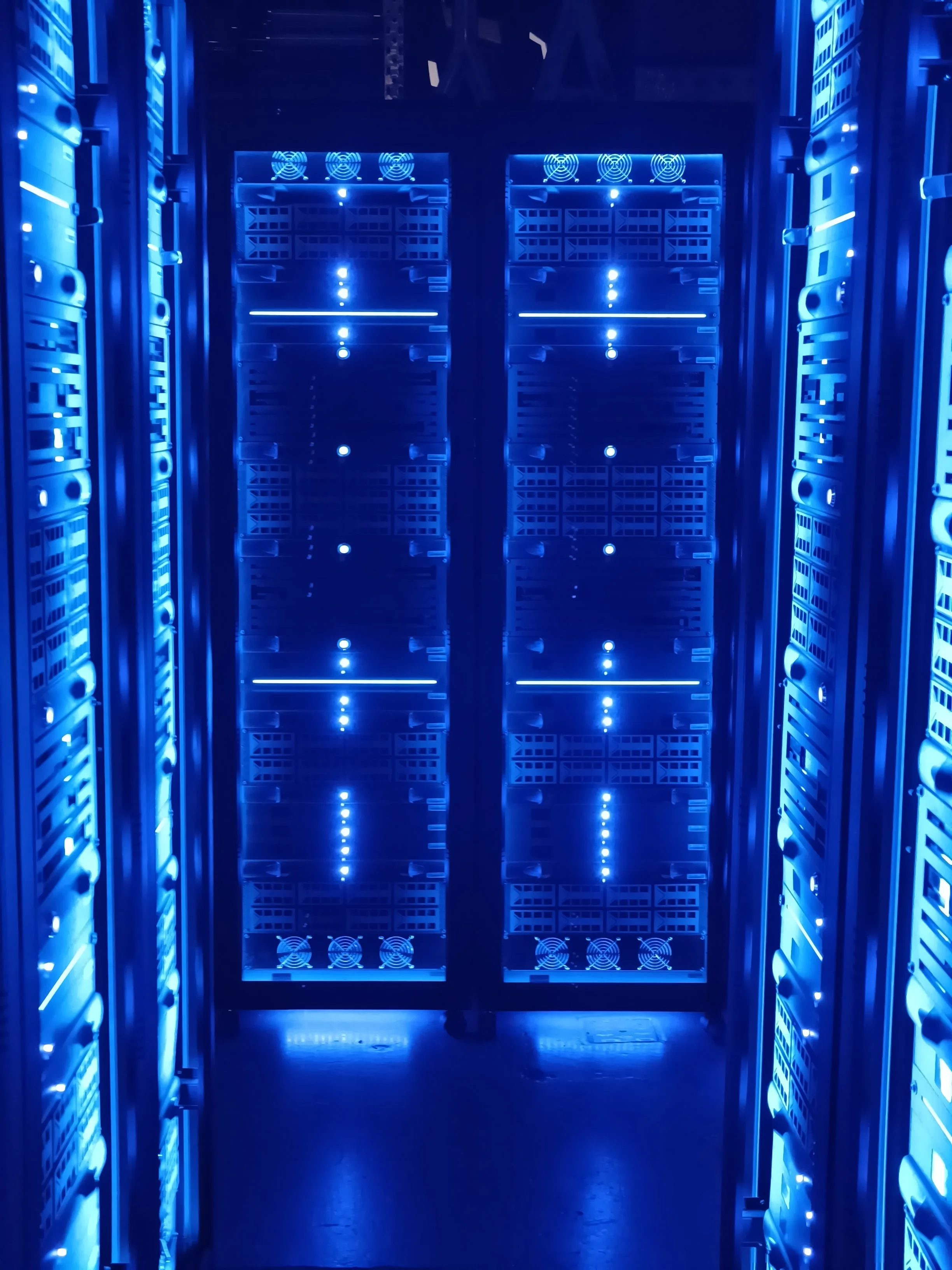 View of a data center with servers illuminated by blue LED lights, and cooling fans at the top and bottom of the server racks.