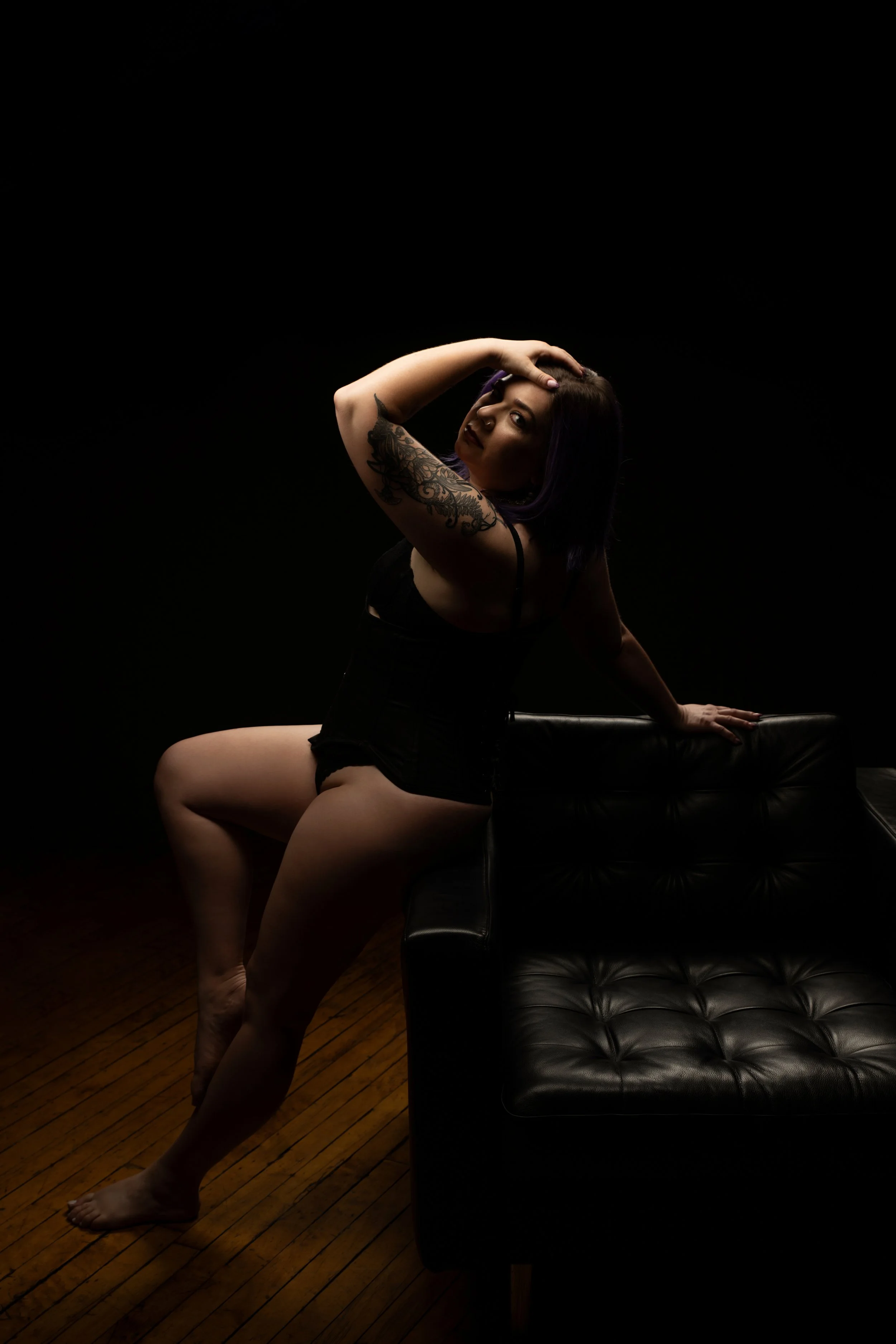 A woman with a tattoo on her arm poses on a black leather couch in a dark room, with low lighting highlighting her features.