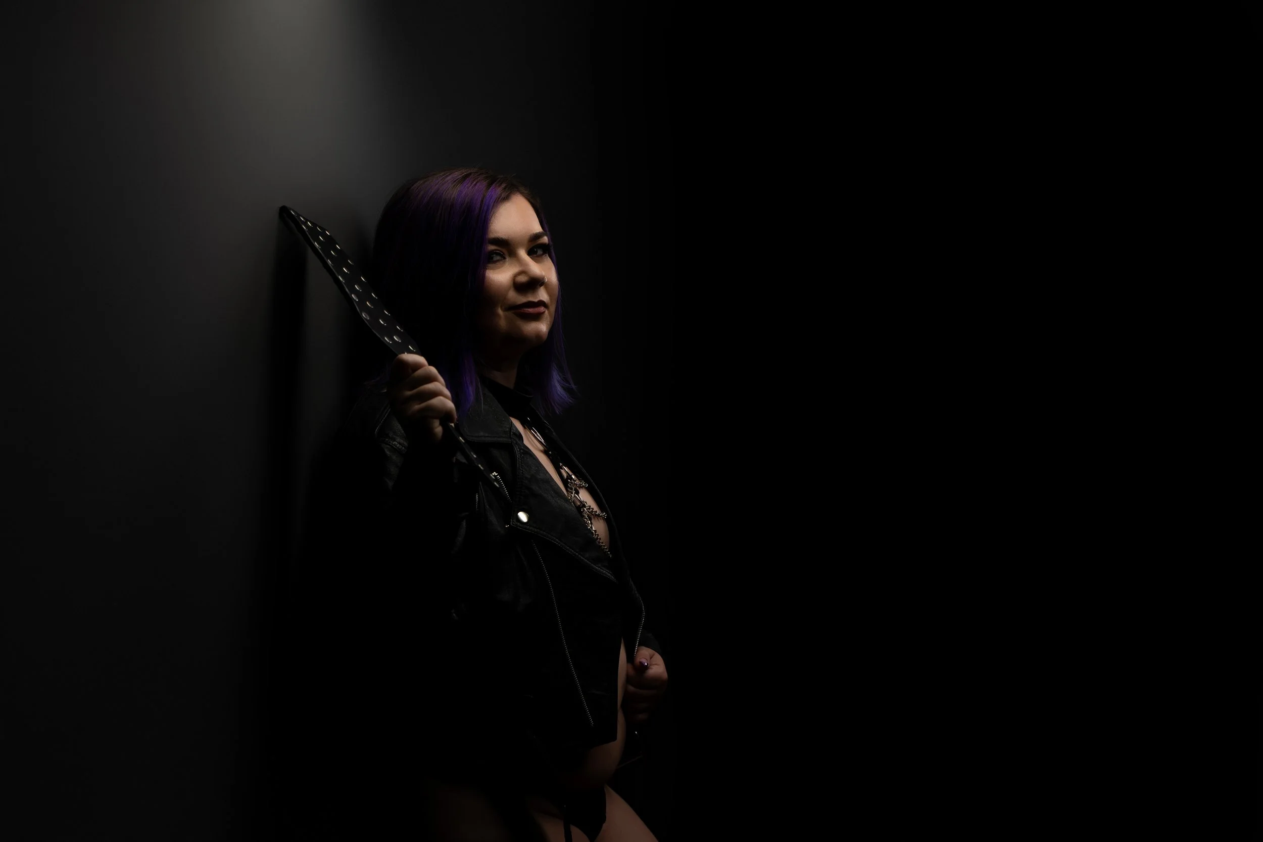 A woman with purple hair holding a knife, wearing a leather jacket and dark clothing, stands against a dark background with minimal lighting.