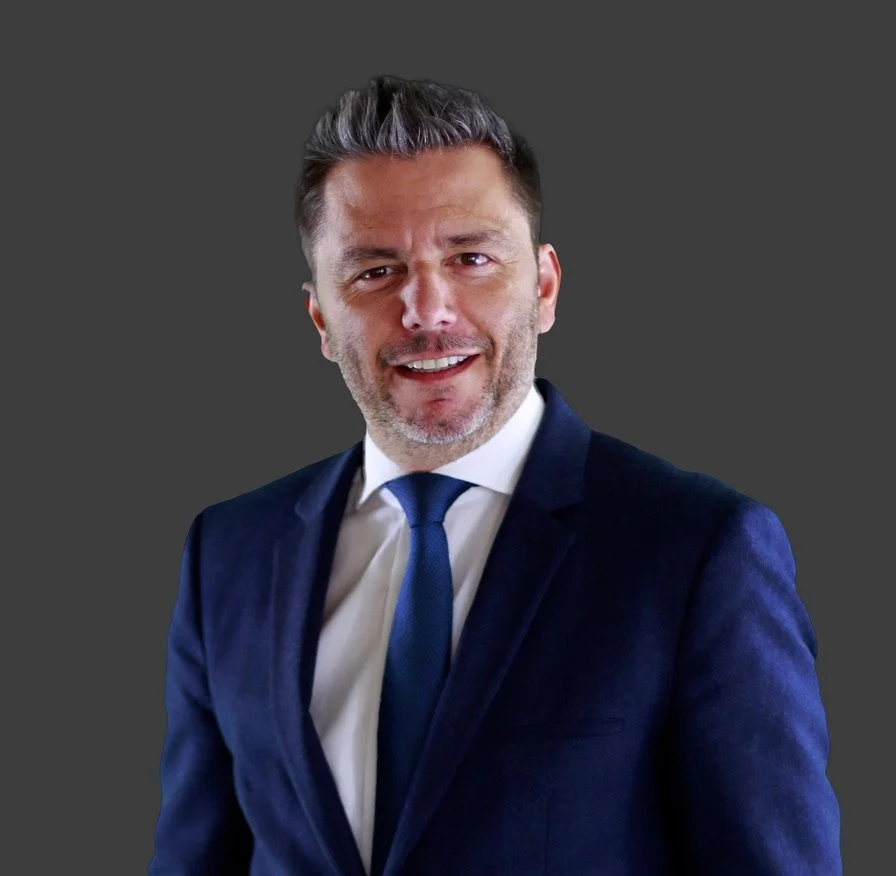 Un homme souriant, portant un costume sombre et une cravate bleue, contre un fond gris.