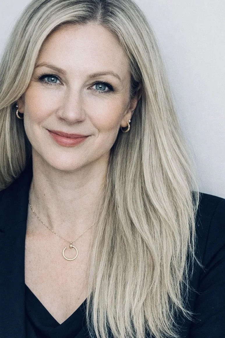 Close-up of a smiling blonde woman with blue eyes, wearing gold hoop earrings, a gold necklace with a circular pendant, and a dark blazer.