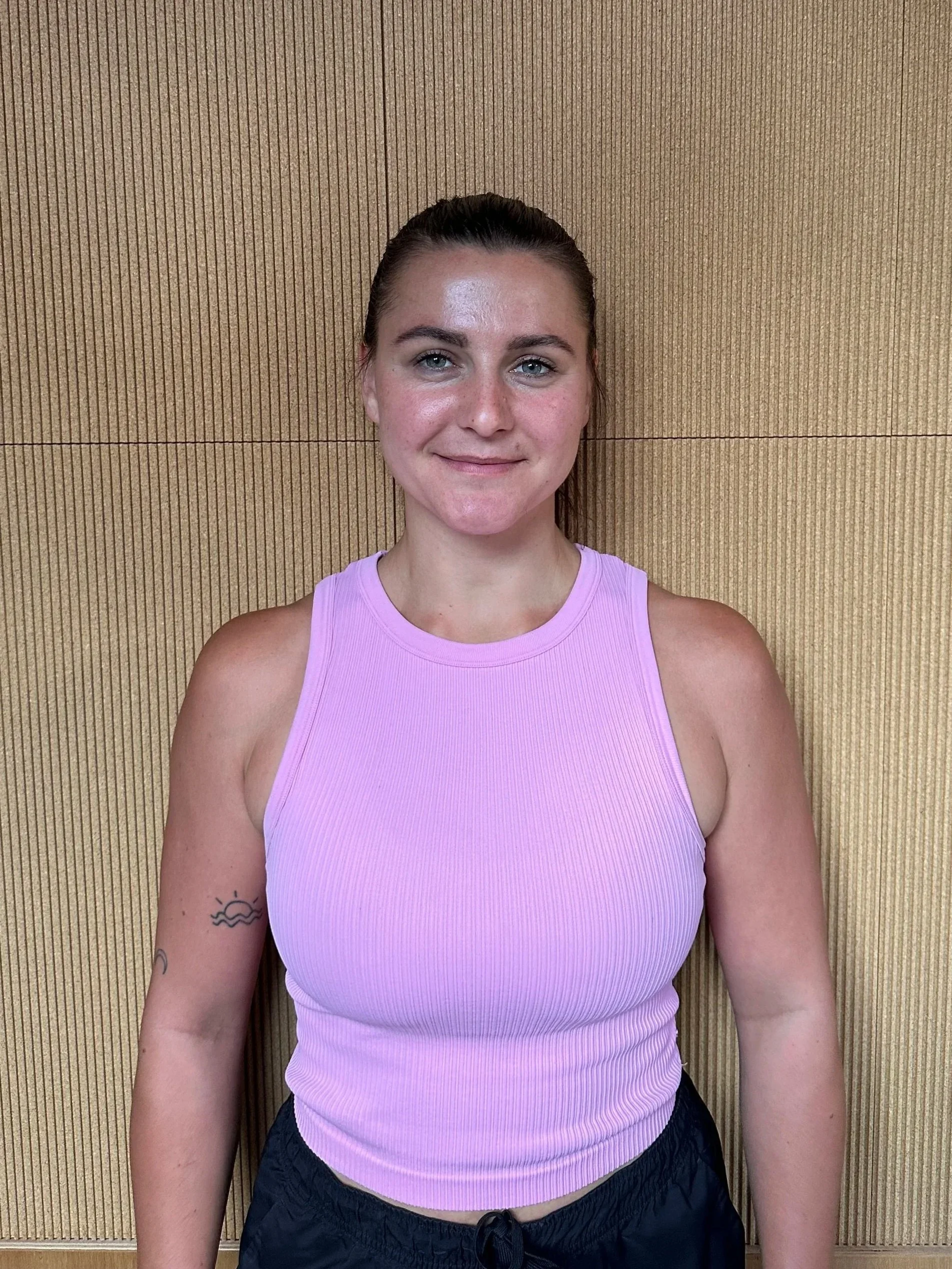 Close-up of a smiling woman wearing a pink sleeveless top, standing against a wooden wall background.