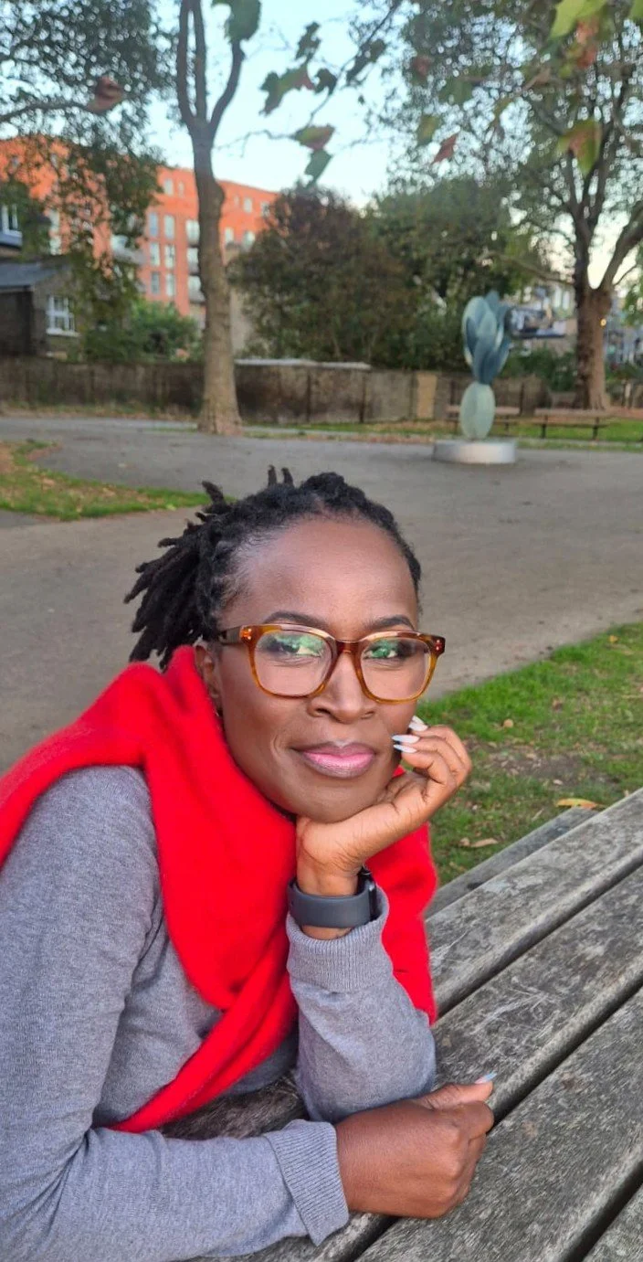 A woman with glasses and dreadlocks resting her chin on her hand sitting on a park bench with trees, a sculpture, and buildings in the background.