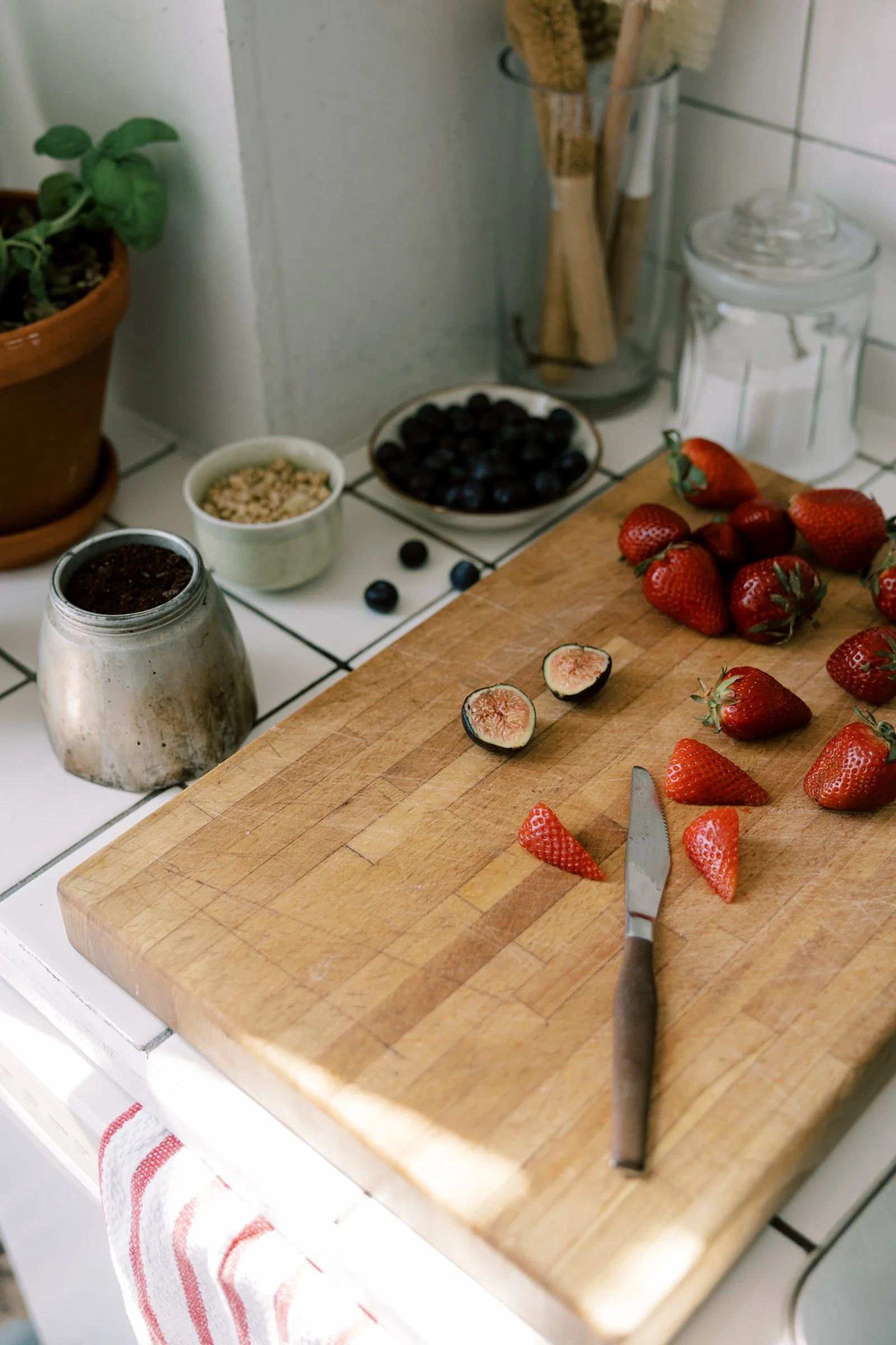 A wooden cutting board with sliced strawberries and a knife, surrounded by whole strawberries, figs, blueberries, and other ingredients on a kitchen countertop.