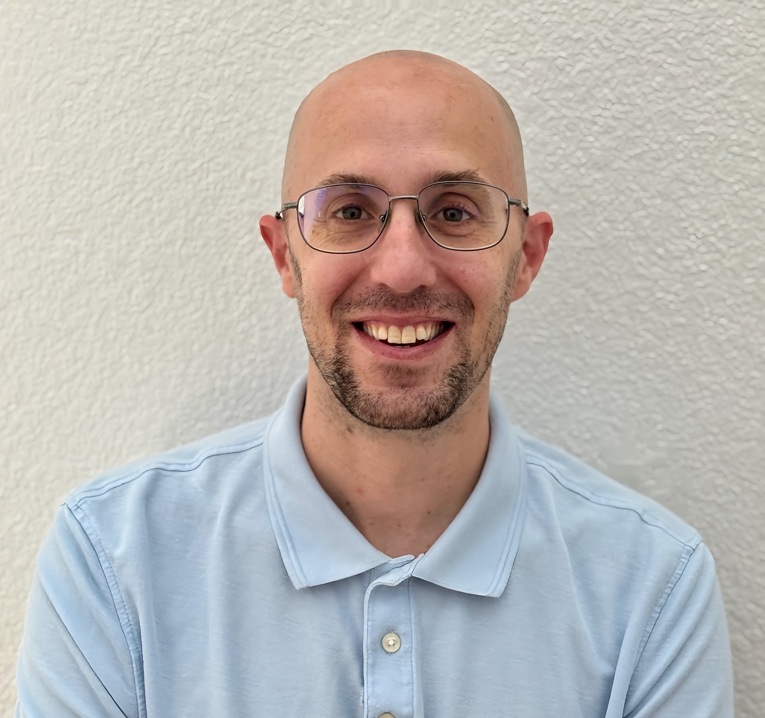 A man with glasses and a beard, smiling, wearing a light blue collared shirt, standing against a textured white wall.