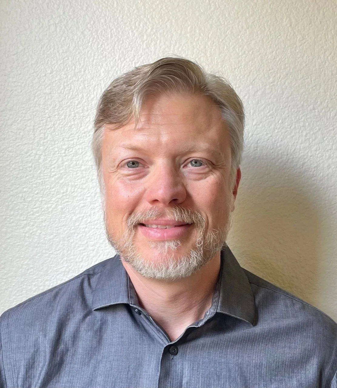 A middle-aged man with gray hair, a beard, and blue eyes, smiling, wearing a dark gray collared shirt, stands against a light-colored textured wall.