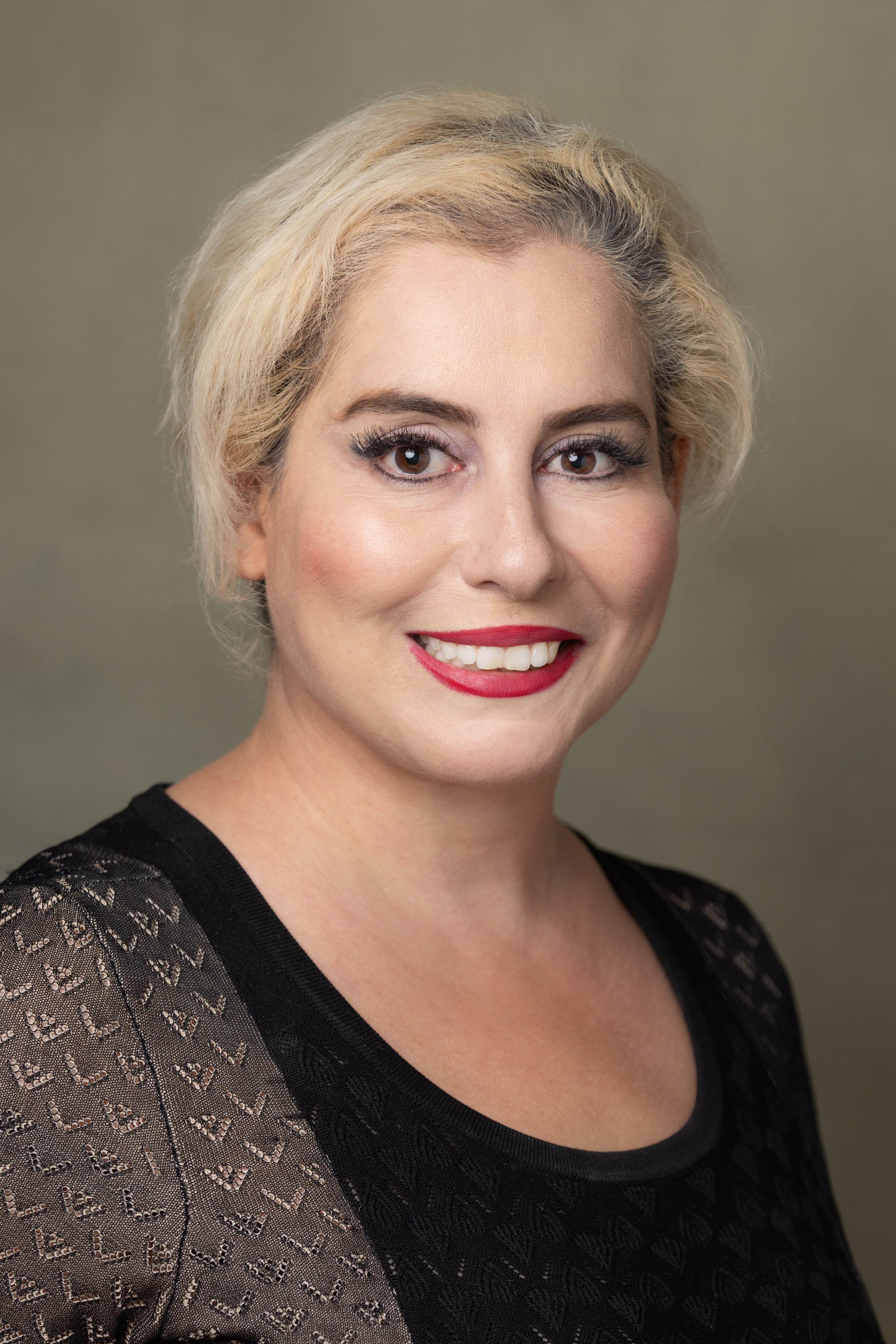 Portrait of a woman with blonde hair, red lipstick, and a black lace top, smiling against a neutral background.