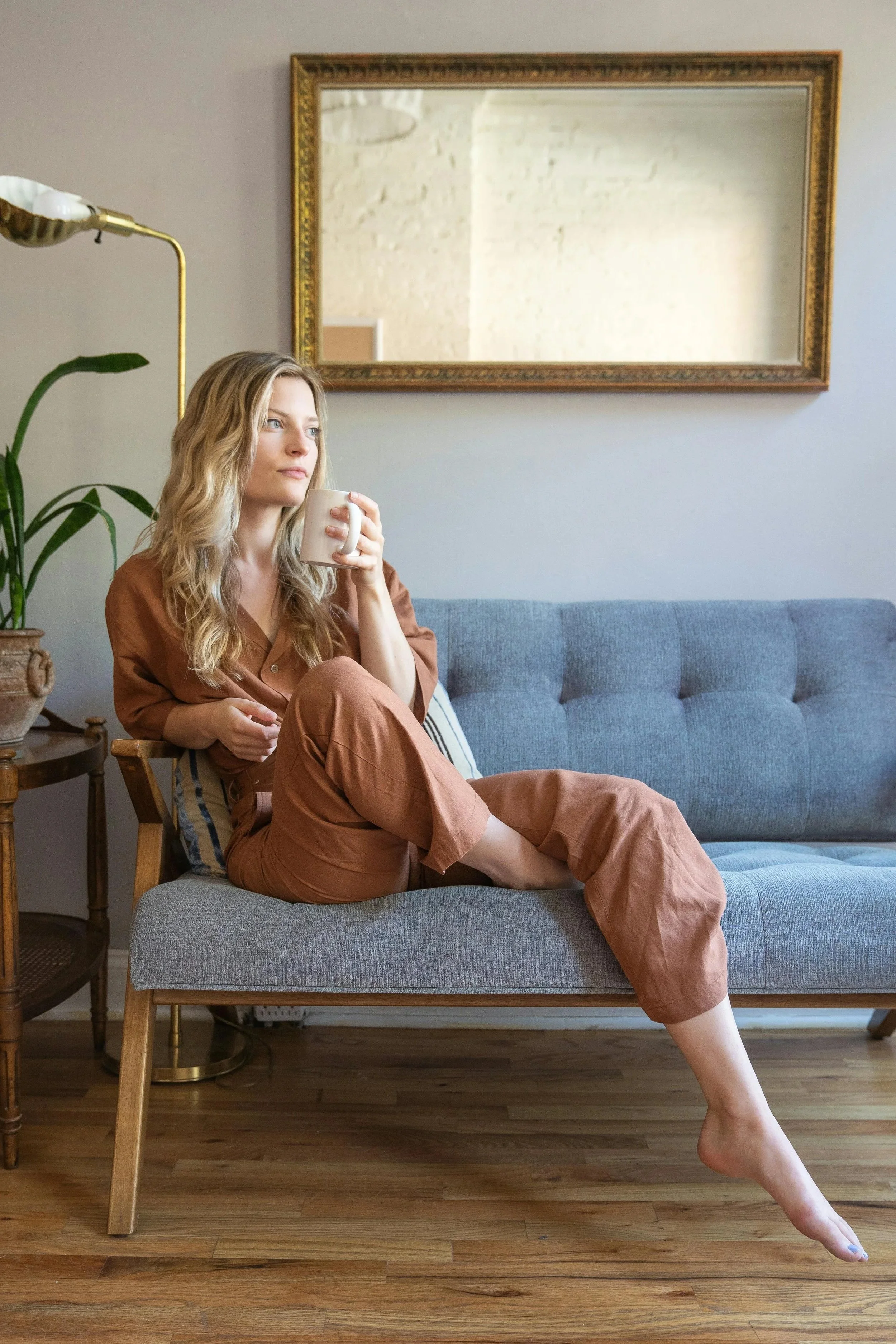 A woman with wavy blonde hair sitting on a gray couch, holding a coffee mug, looking pensively to the side in a cozy living room with a large mirror on the wall, a houseplant, and wooden flooring.